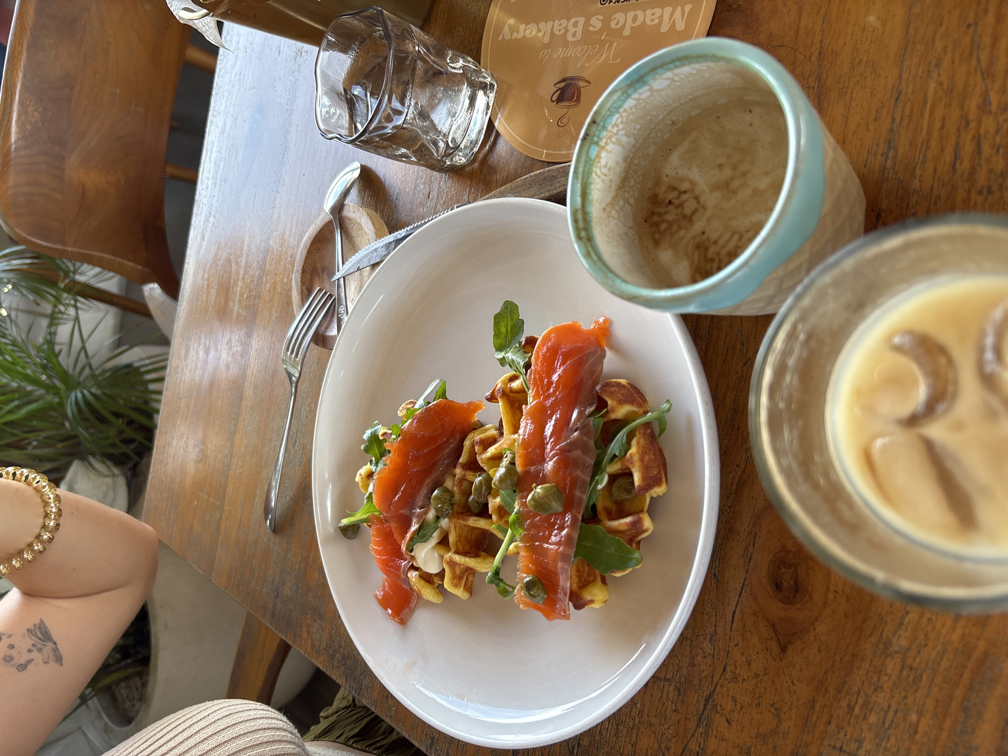 The image captures an overhead view of a table set for a meal, likely breakfast or brunch, at a cafe in Kuta Selatan, Indonesia.
The primary subject is a white plate holding savory waffles topped with slices of smoked salmon, fresh arugula, numerous capers, and a small dollop of white sauce. To the right of the plate, there's an empty ceramic mug with a light blue rim and a clear glass containing an iced coffee. An empty, textured clear glass and metal cutlery (a fork, knife, and spoon) rest on a small wooden coaster to the left of the plate.
Partially visible in the bottom left is the arm of a person, adorned with a golden beaded bracelet and a small tattoo of a winged figure with stars. A wooden chair and green potted plants are also visible in the background, hinting at the cafe's decor. The table itself is made of light-colored wood.
The activity taking place is dining, with a focus on the food and beverages. The bright, natural lighting suggests it is daytime, likely morning or early afternoon.
A notable detail is the text "Welcome to Made's Bakery" printed on the wooden coaster, identifying the establishment. The overall scene conveys a relaxed and inviting atmosphere.