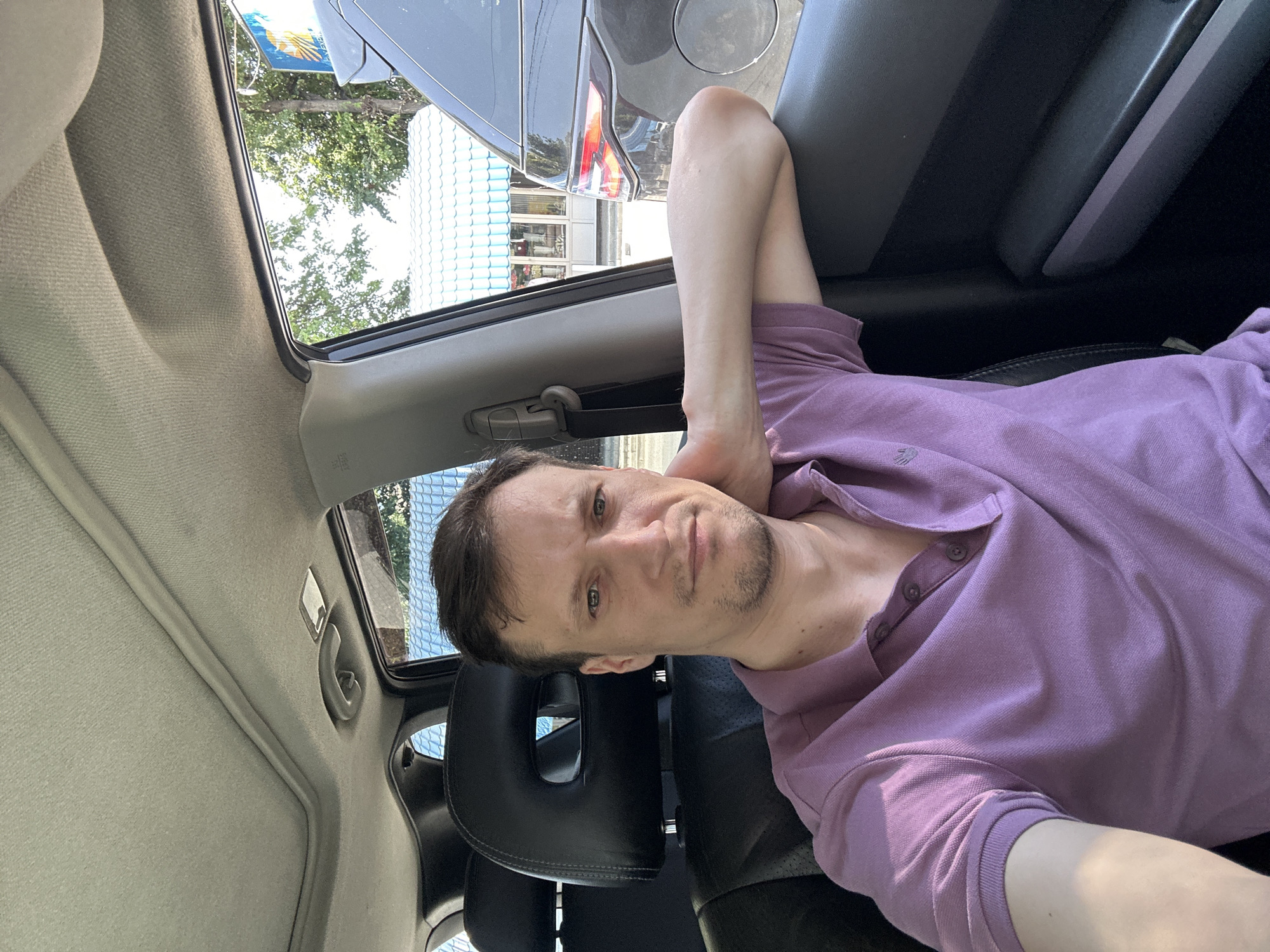 The image shows a man sitting in the driver's seat of a car, taken from a selfie perspective. He is wearing a light purple polo shirt and has his right arm resting on the open car door. He is looking towards the camera with a neutral expression. Through the car window, a dark-colored SUV is visible, stopped in traffic behind the man's car. In the background, there are buildings with light blue tiled roofs and green trees. The scene appears to be in an urban or suburban area, likely during daylight hours, given the natural lighting. The specific location of Kamianets-Podilskyi, Ukraine is not discernible from visual cues within the image.