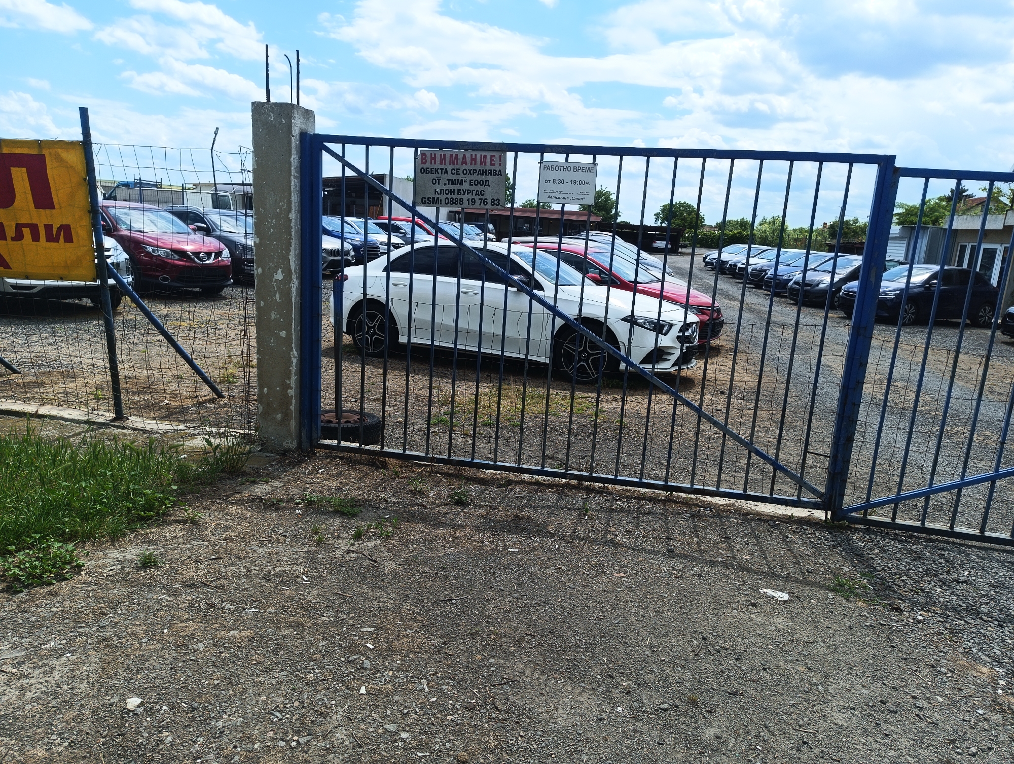 The image shows an outdoor car lot. Cars of various makes and models are visible behind a blue metal fence. A sign on the gate indicates the lot is located in Burgas, Bulgaria and is secured by "TIM" EOOD. The operating hours are listed as 8:30-19:00. The ground is gravel. Overcast skies are visible.