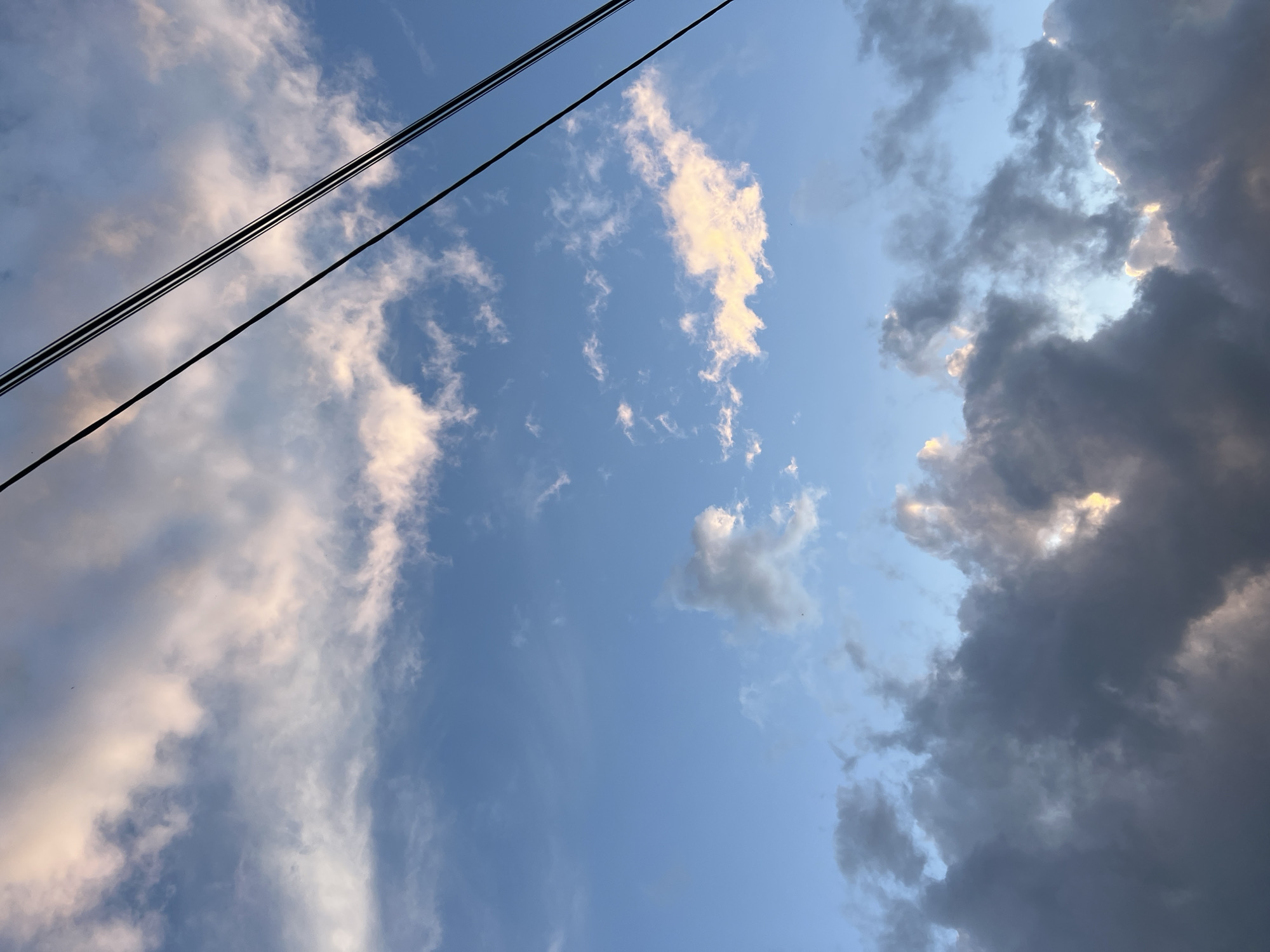 The image shows a sky with varying cloud formations. On the left side, there are lighter, white clouds with a touch of color suggesting either sunrise or sunset. The central area presents a clear blue sky with sparse, small clouds. On the right side, darker, stormier clouds are gathered, hinting at possible rain or a change in weather. Several electrical wires are visible in the upper-left corner, running diagonally across the frame. There is no information to suggest the location is in Chaikovsky, Russia, or any other place. No people, actions, or interactions are visible.