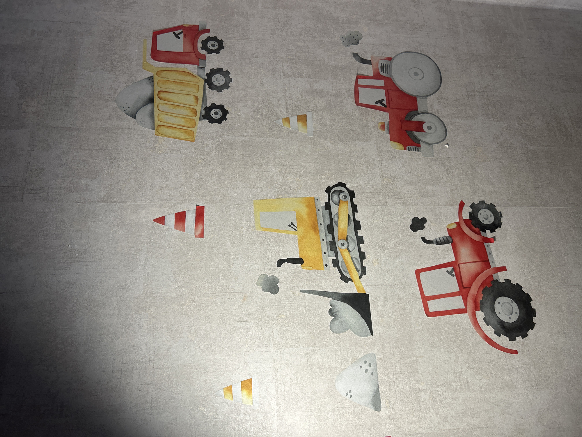 The image displays a wall covered in a textured, light-colored wallpaper. Adorning the wall are several cartoon-style decals of construction vehicles and traffic cones. In the upper right quadrant, a red dump truck filled with gray rocks is depicted. To its left, a red and white striped traffic cone stands. In the center of the image, a yellow bulldozer is shown with its front loader raised, a pile of gray sand to its left, and a puff of black smoke emanating from its exhaust. Below and to the right of the bulldozer, another red and white traffic cone is visible. In the lower right quadrant, a red tractor with gray wheels is parked, also emitting a puff of black smoke. In the lower left quadrant, another red tractor is positioned, this one with large black wheels and a smaller gray wheel at the rear. The scene is devoid of people or any indication of a specific location in Saint Petersburg, Russia. The lighting suggests a relatively dim indoor environment, with a gradient of light and shadow across the wallpaper.