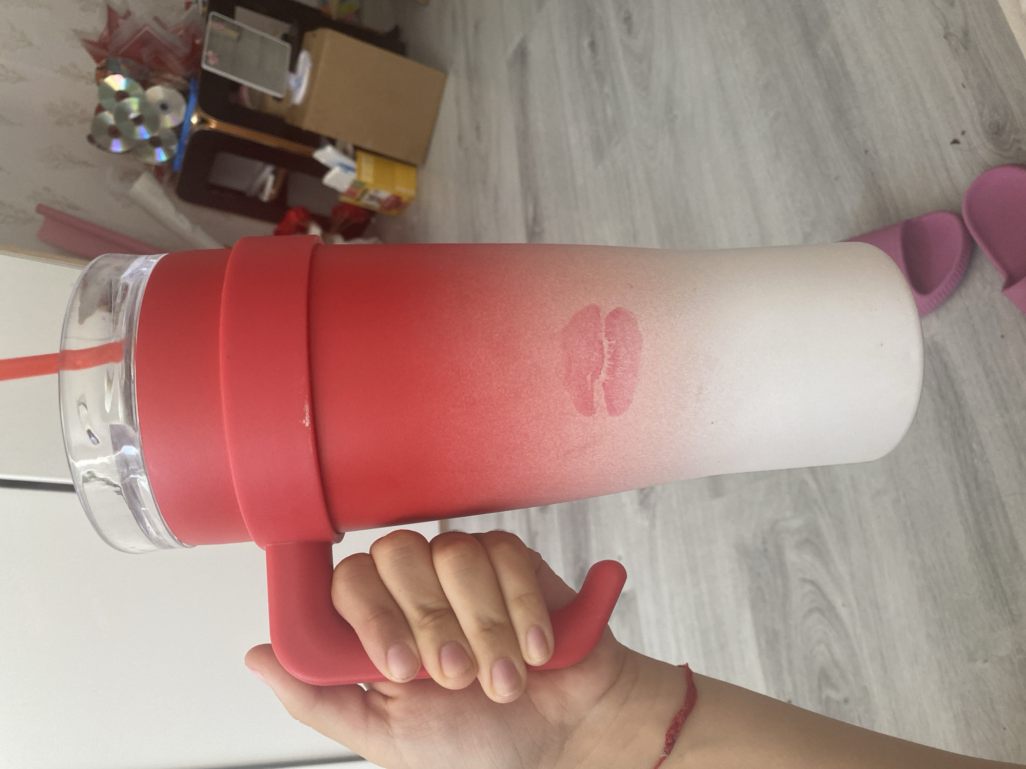A person's hand is holding a red and white gradient tumbler with a red handle. A lipstick imprint of lips is visible on the tumbler. The tumbler has a clear lid and a red straw. In the background, there are some shelves with various items, including CDs, and pink slippers on the floor. The environment appears to be indoors, possibly a room. There is no information in the image to suggest this location is in Blagoevgrad, Bulgaria.