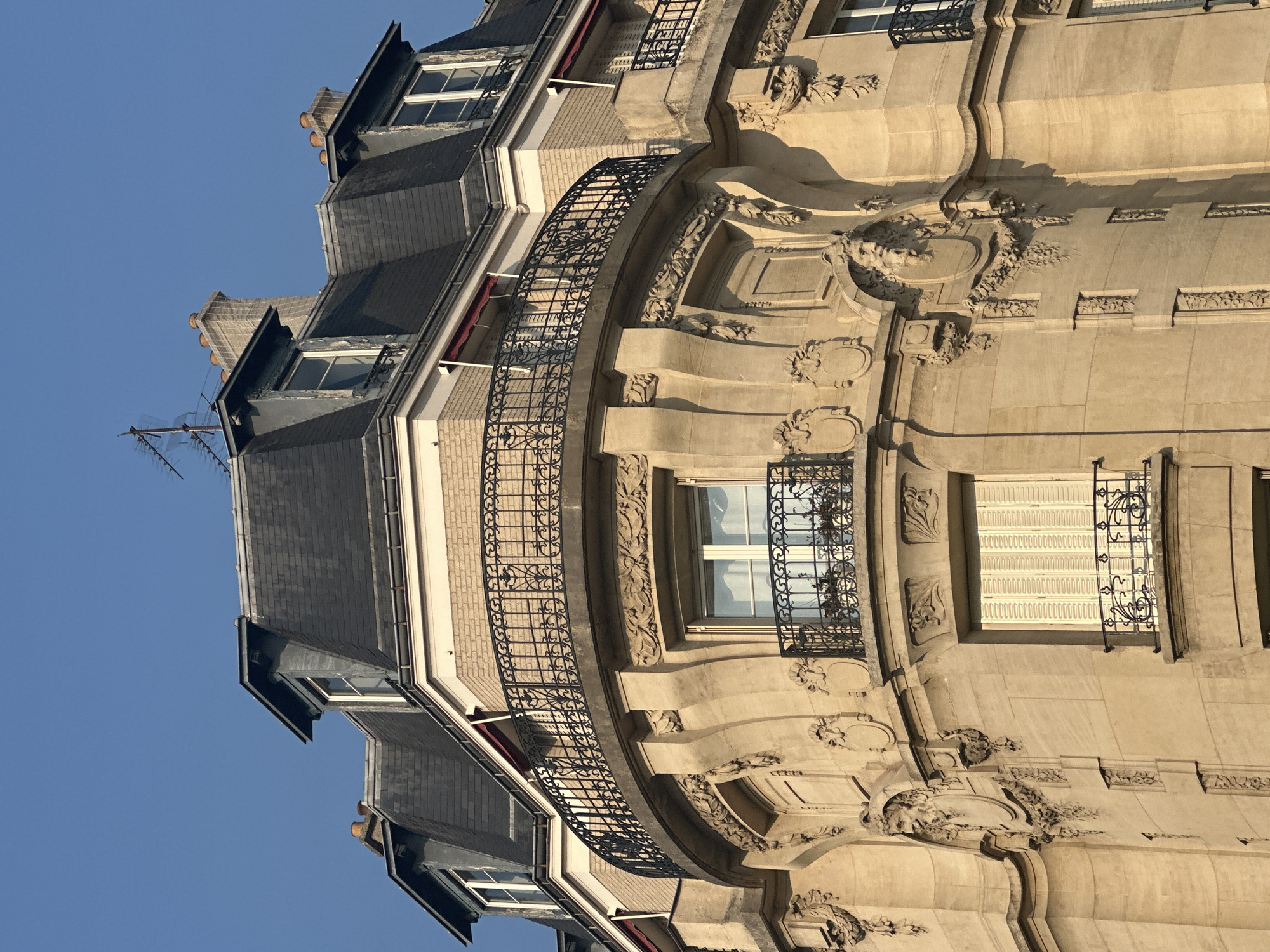 The image shows the upper floors of a Parisian Haussmannian building on a clear day. The building features a mansard roof with dark grey slate tiles and dormer windows. The facade is constructed of light-colored stone blocks, adorned with ornate carvings and decorative elements, including a prominent curved balcony with wrought-iron railings on the upper floor. There are multiple windows visible, some with closed shutters and small wrought-iron balconies. The architecture is typical of the late 19th and early 20th centuries, common in central Paris. No people are visible in the image.