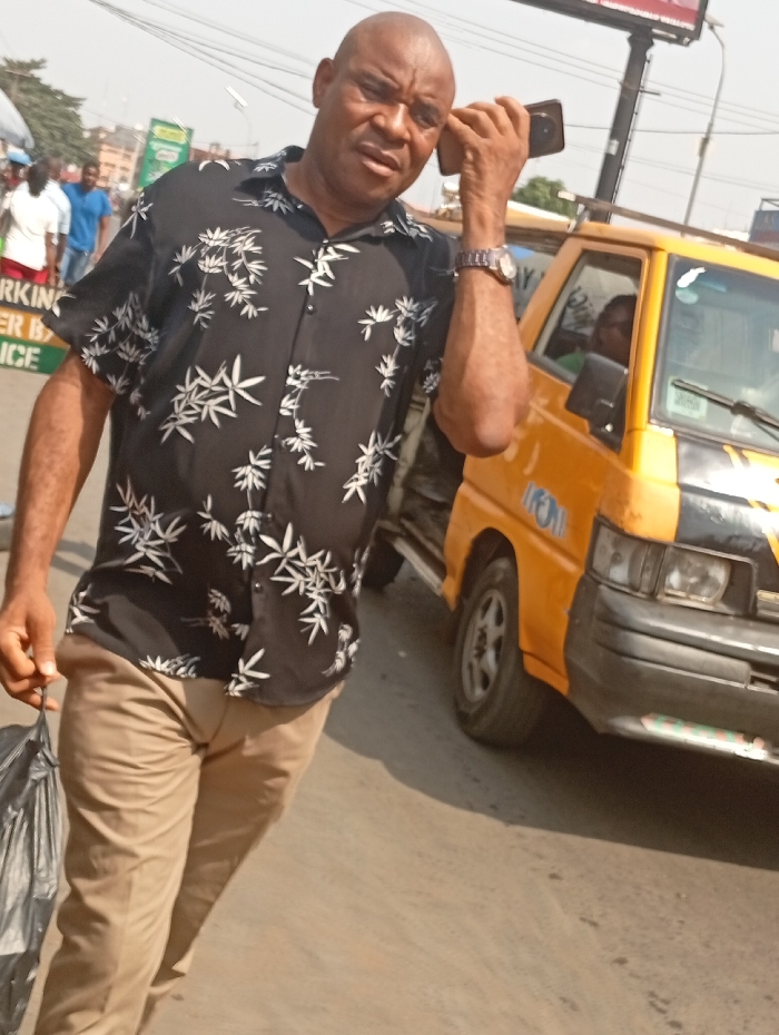 The image depicts an outdoor urban scene in Onitsha, Nigeria, under daylight conditions. A bald, middle-aged male, appearing to be of African descent, is in the foreground, walking towards the viewer. He wears a black short-sleeved button-up shirt with a white bamboo leaf pattern, khaki trousers, and a silver-toned wristwatch on his left wrist. He holds a black smartphone to his right ear with his right hand, and a black plastic bag in his left hand. His gaze is directed towards the camera.
To the right of the main subject, a yellow commercial minibus or taxi, an older model with partial 'TREM' lettering on its side, is visible on the paved road. A person's face is partially discernible through the vehicle's driver-side window. In the left background, several blurred individuals are present on what appears to be a sidewalk, including one person in a white shirt and another in a blue shirt. A partially visible sign in the far left background reads "PARKING...BY POLICE." Overhead cables and a billboard structure are visible in the upper background. The ground is paved, appearing light grey or tan.