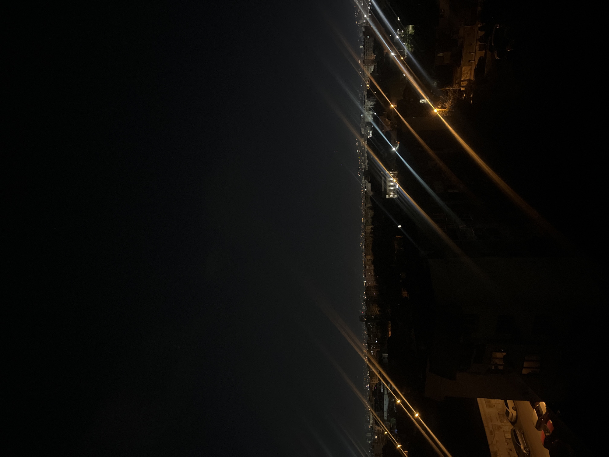 The image depicts a nighttime scene, likely overlooking a city from a high vantage point. The left side of the image is dominated by a large dark area, potentially representing a body of water or a dark expanse. The right side of the image shows a line of lights stretching into the distance, likely streetlights and building lights along a coastline or a major road. The lights appear as streaks due to a long exposure or movement during the photograph. Specific details about Sofia, Bulgaria, are not evident in the image without additional context or recognizable landmarks.