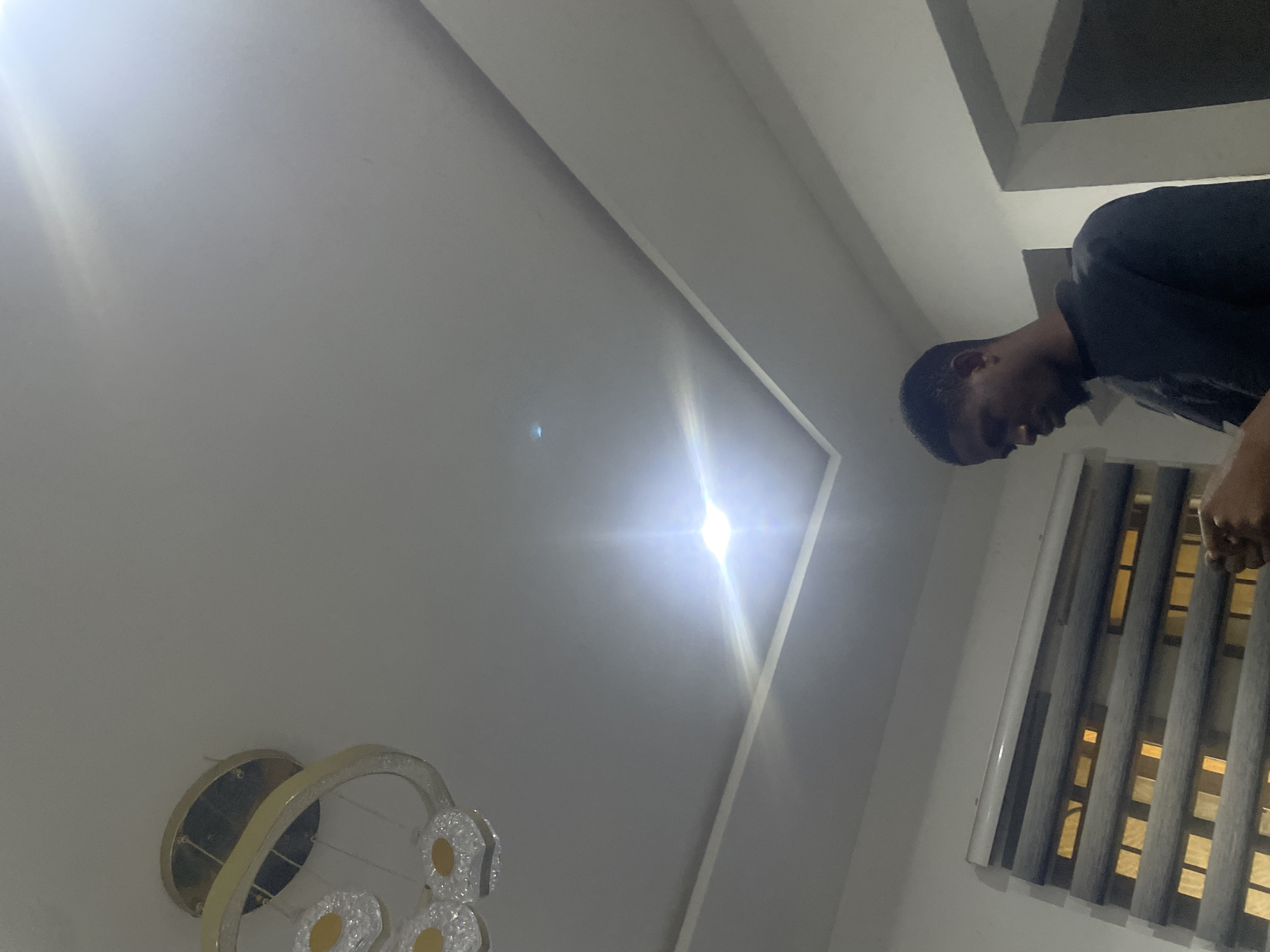 In a room with a recessed ceiling, a young Black male stands facing downwards, looking at his phone which he holds in his right hand. He is wearing a dark t-shirt. To his left, a window is covered by horizontal blinds, with lights visible through them. A modern chandelier with circular elements is mounted on the ceiling in the upper left corner of the room. The room is illuminated by a bright light fixture in the center of the recessed ceiling. The overall impression is of an indoor, domestic setting. The image may have been taken in Abuja, Nigeria, based on the prompt's contextual information.