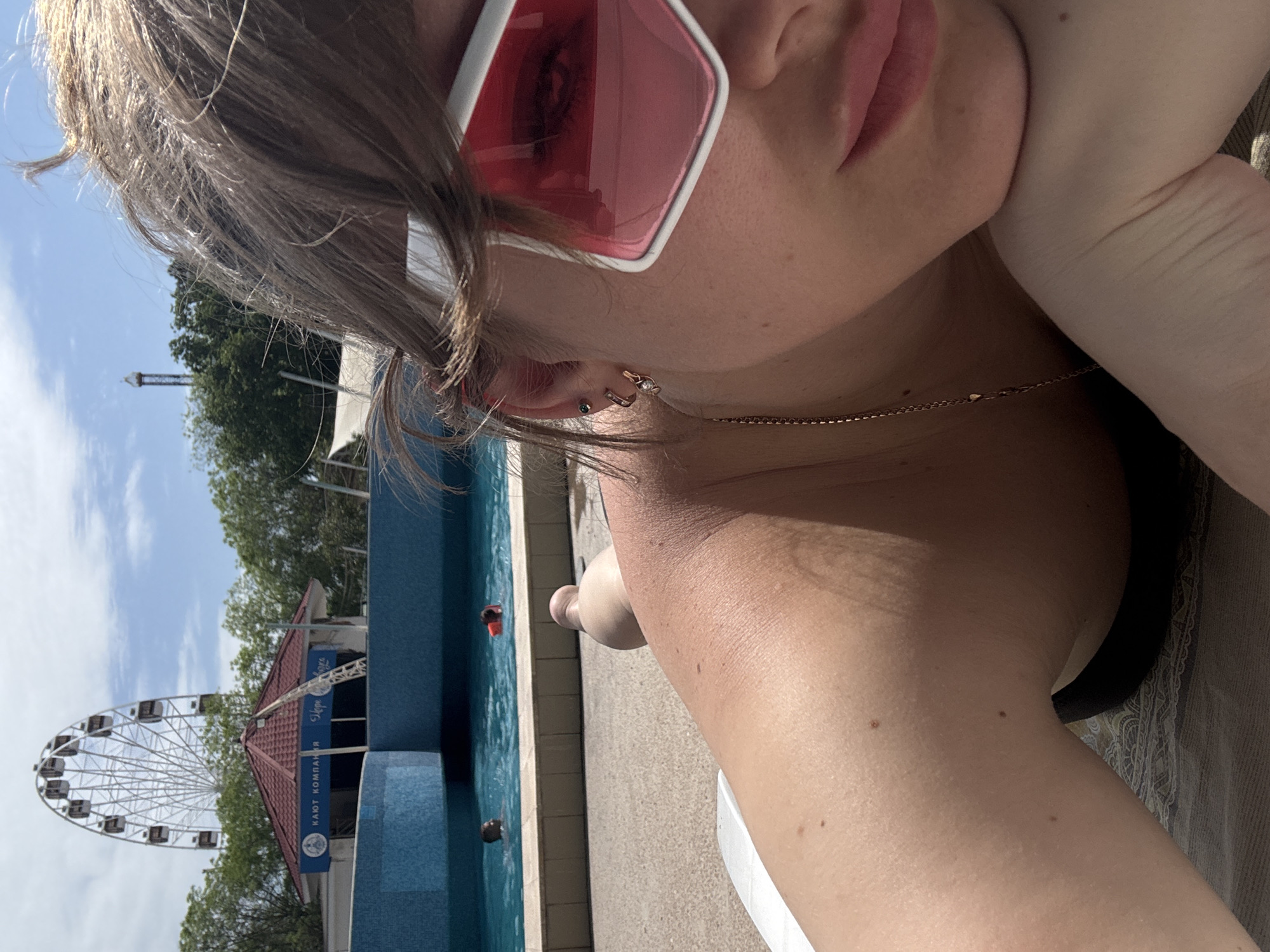 A fair-skinned person wearing pink-tinted glasses and earrings is reclining near a swimming pool. A gold chain is visible around their neck. In the background is a Ferris wheel, a building, and a tall tower against a blue sky with clouds. People are visible in the pool. Based on the Ferris wheel and architecture, the location is likely in Yevpatoriya, Ukraine.