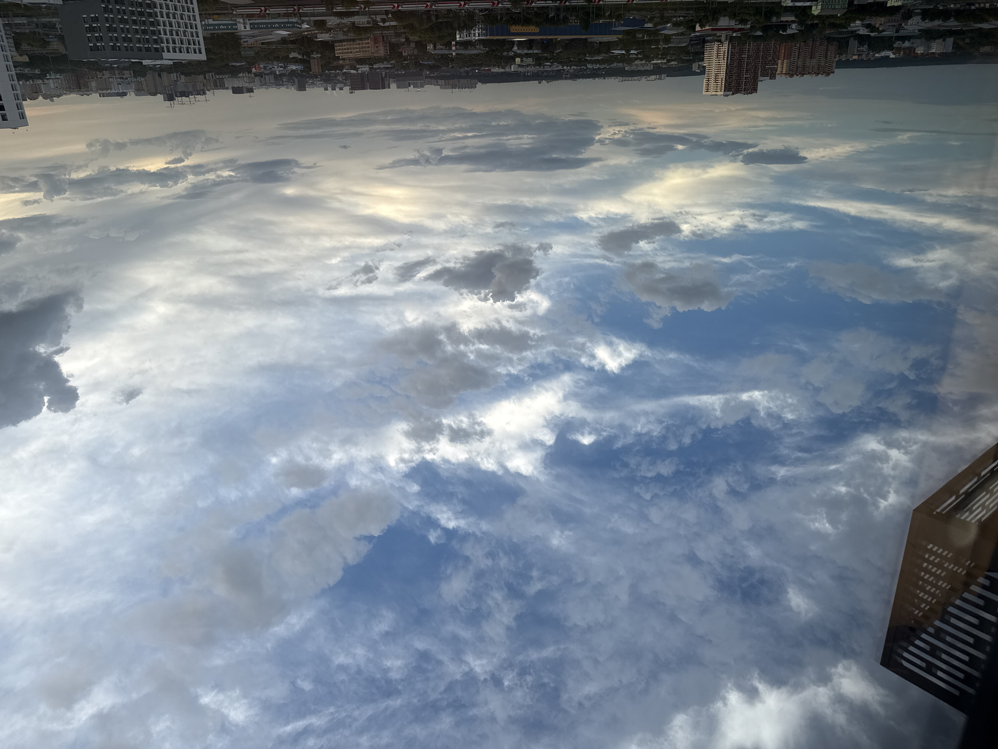 A wide-angle, eye-level shot captures a view of a city skyline under a dramatic sky, taken from an elevated position in Lyubertsy, Russia. The upper portion of the frame is dominated by a complex cloudscape, with streaks of white and grey clouds interspersed with patches of blue sky. The clouds vary in density, with some appearing thin and wispy, while others are thicker and more textured. The lighting suggests it is either dawn or dusk, with a warm glow visible on the horizon.
In the foreground, a dark, angular structure with horizontal slats partially obscures the left side of the image, likely a building element or balcony.
The lower part of the image reveals a cityscape featuring a dense collection of buildings. Numerous apartment blocks of varying heights and architectural styles are visible, some with brick facades and others with lighter-colored exteriors. Trees are scattered throughout the urban landscape, and some construction cranes are present, indicating ongoing development. A train track with a red and white train can be seen in the lower foreground. The overall impression is of a bustling, modern urban environment viewed from a distance.