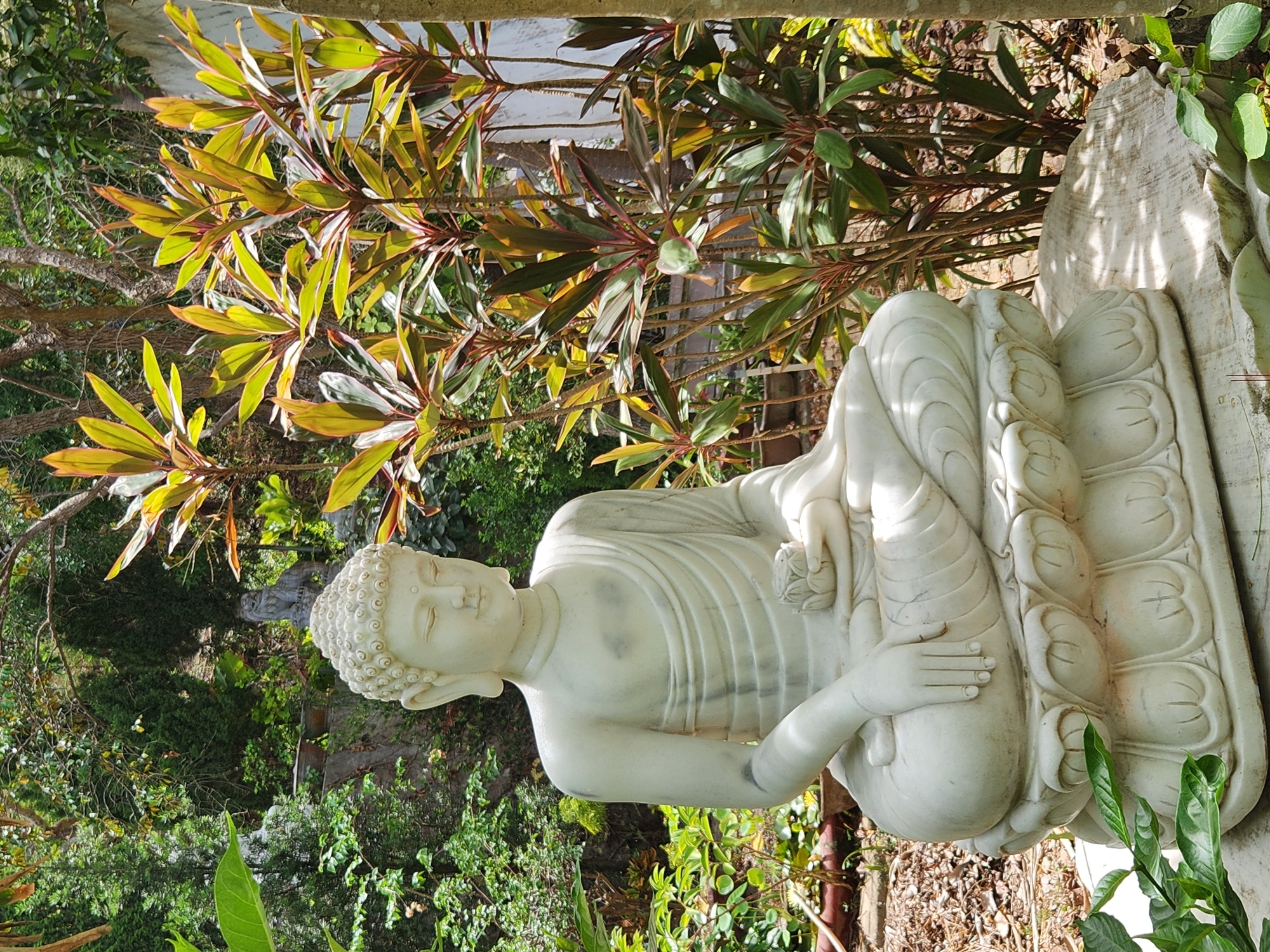 A white marble statue of Buddha sits in a lotus position outdoors, holding a lotus flower in its left hand. The statue is on a carved lotus base, which rests on a large, rough stone. The background features lush greenery with various plants and trees, suggesting a garden or natural setting. Sunlight filters through the foliage, creating dappled light and shadows. The overall scene conveys a sense of peace and tranquility. The location is the Marble Mountains in Da Nang, Vietnam, a famous Buddhist pilgrimage site known for its caves, temples, and stunning natural beauty.