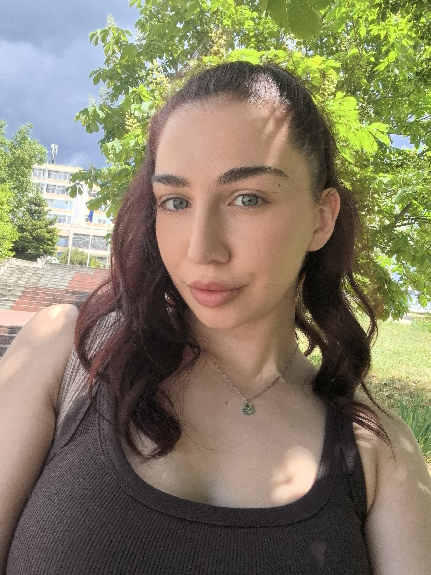 A woman takes a selfie outdoors on a sunny day. She has fair skin, light eyes, and auburn hair pulled back. She wears a brown tank top and a silver necklace with a heart-shaped pendant. Behind her, there's a tree with green leaves and a building with stairs leading up to it. The sky is partly cloudy. The exact city and location are unknown.