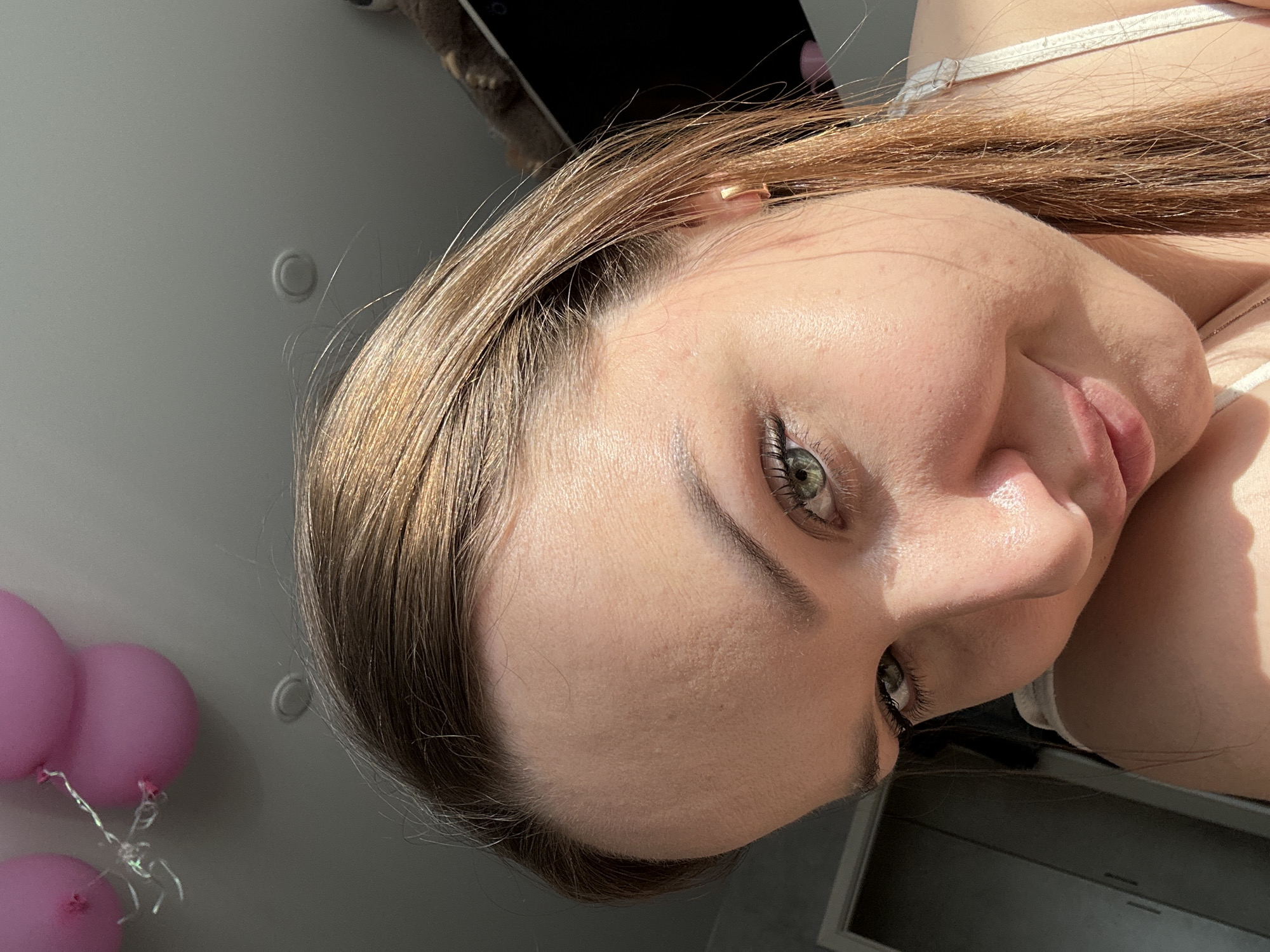 The image displays a close-up, high-angle portrait of a female individual. She possesses light brown hair with reddish undertones, styled to fall over her right shoulder. Her eyes are green, and she is looking directly at the camera with a slight, closed-mouth smile. Her face is partially illuminated by direct sunlight, creating highlights on her right cheek and forehead. A small gold hoop earring is visible in her right earlobe. A white strap, consistent with a top or camisole, is visible on her right shoulder. In the upper left background, three pink balloons tied with shimmering silver string are attached near a light grey or white ceiling. A small, circular, raised fixture is also present on the ceiling. To the upper right, a dark rectangular object, possibly a screen or electronic device, is partially visible, along with a dark, furry object above it. To the lower left, a light-colored door frame and a dark vertical panel, possibly a wardrobe or cupboard, are discernible. The specific location within the city of Chaikovsky, Russia, is not discernible from the visual information provided.