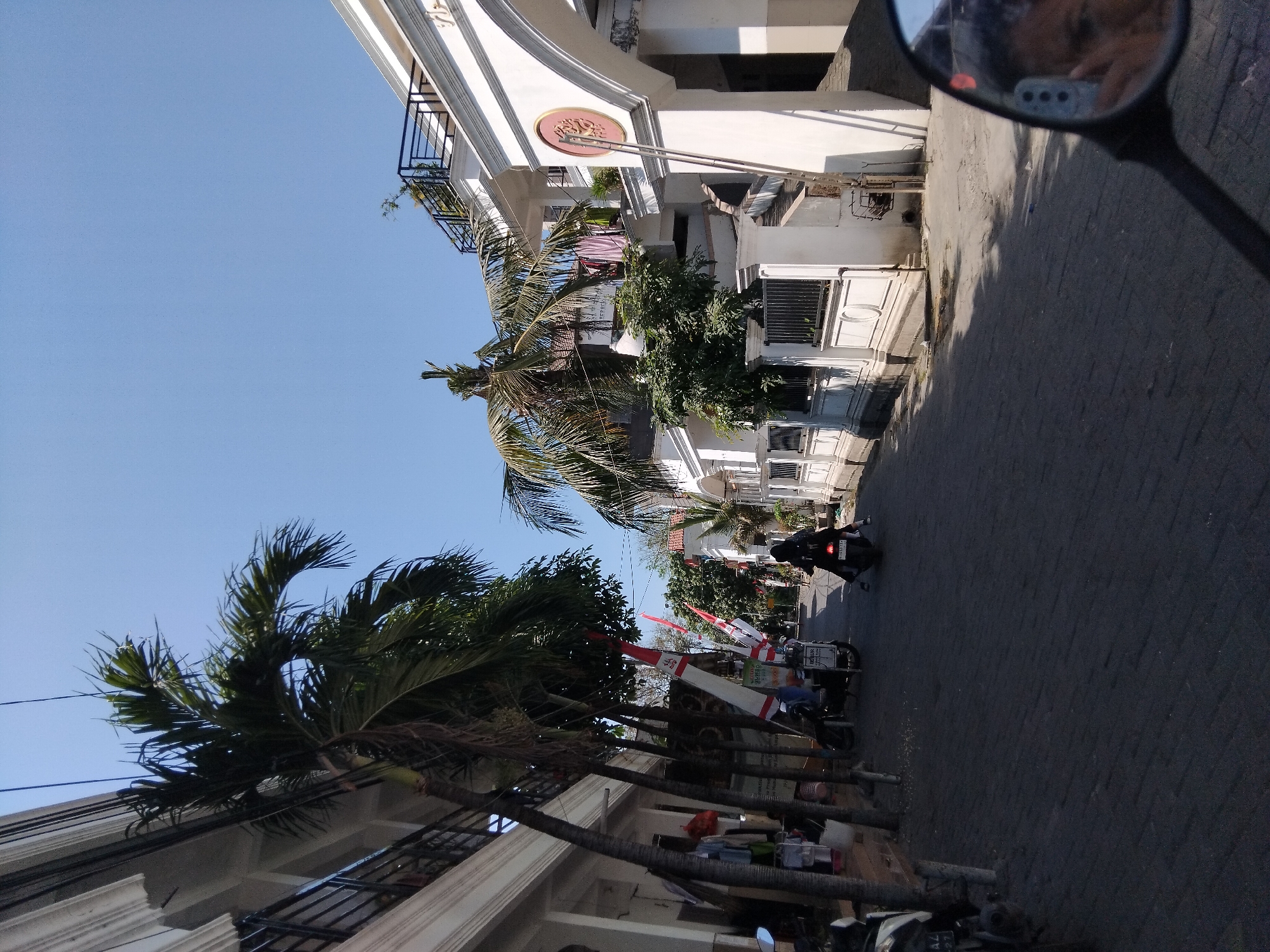 The image depicts a street scene in Berbek, Indonesia, under a clear blue sky. The perspective appears to be from a motorcycle, as evidenced by the reflection of a person's face and hand in the motorcycle's rearview mirror, which is in the foreground on the right. The street is paved with dark grey interlocking bricks. On the left side of the street, there is a white building with multiple stories, featuring balconies with dark railings. Several tall, slender palm trees with sparse fronds are positioned along the side of this building, with some clothes hanging on lines between them. Indonesian flags, red and white, are mounted on poles along this side of the street.
On the right side of the street, a white building with an arched entrance is prominent. This building also has balconies with dark railings and is decorated with white ornate trim. A tree with dense green foliage is visible in front of this building. Beyond this, further down the street, other white buildings and more greenery can be seen.
A person wearing dark clothing and a headscarf is riding a motorcycle away from the camera down the street. A few other motorcycles are parked or in motion further down the road. The overall atmosphere suggests a typical day in a residential or commercial area within the city, with a mix of modern architecture and tropical vegetation.