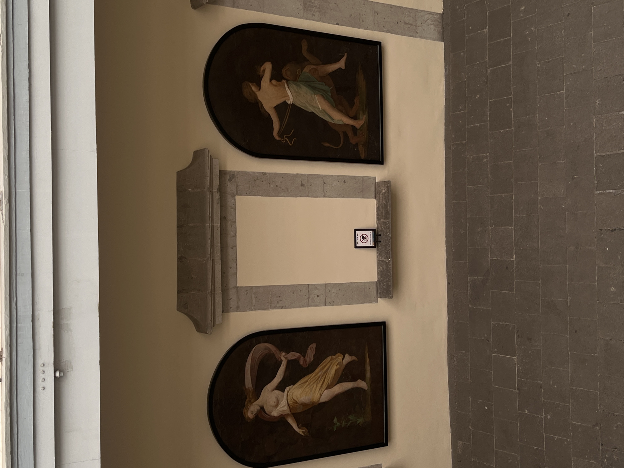 The image shows an indoor corridor or gallery in Mexico City, Mexico, featuring two framed paintings set within architectural niches on a light-colored wall. The perspective is slightly angled, looking down the length of the space, with the image rotated sideways.
The top painting, within an arched black frame, depicts two figures. One figure, mostly nude with a light green drape around its waist, has fair skin and appears to be in an active or struggling pose, holding a rope-like object. This figure interacts with a darker, muscular, and more beast-like figure with horns and hooves, suggesting a mythological scene, possibly a satyr or demon.
Below this, the bottom painting, also in a similar arched black frame, features a fair-skinned female figure, partially draped in flowing yellow and pink fabric. She appears to be floating or reclining dynamically against a dark background, possibly a nymph or goddess, rendered in a classical style.
Between the two paintings, a small sign hangs on the wall, displaying the text "NO TOUCH" prominently, and likely "NO PHOTOS" or "NO FLASH" in smaller text below it, indicating a museum or exhibition setting. The paintings are flanked by decorative stone architectural elements, resembling pilasters or columns.
To the left, a light-colored wall or pillar with gray trim runs vertically, showing some wear. To the right, a dark, rectangular stone-tiled floor extends, providing a clear contrast with the smooth wall. The lighting is even, suggesting an indoor environment, possibly illuminated by artificial lights or indirect natural light. The overall impression is that of a historical building or museum.
