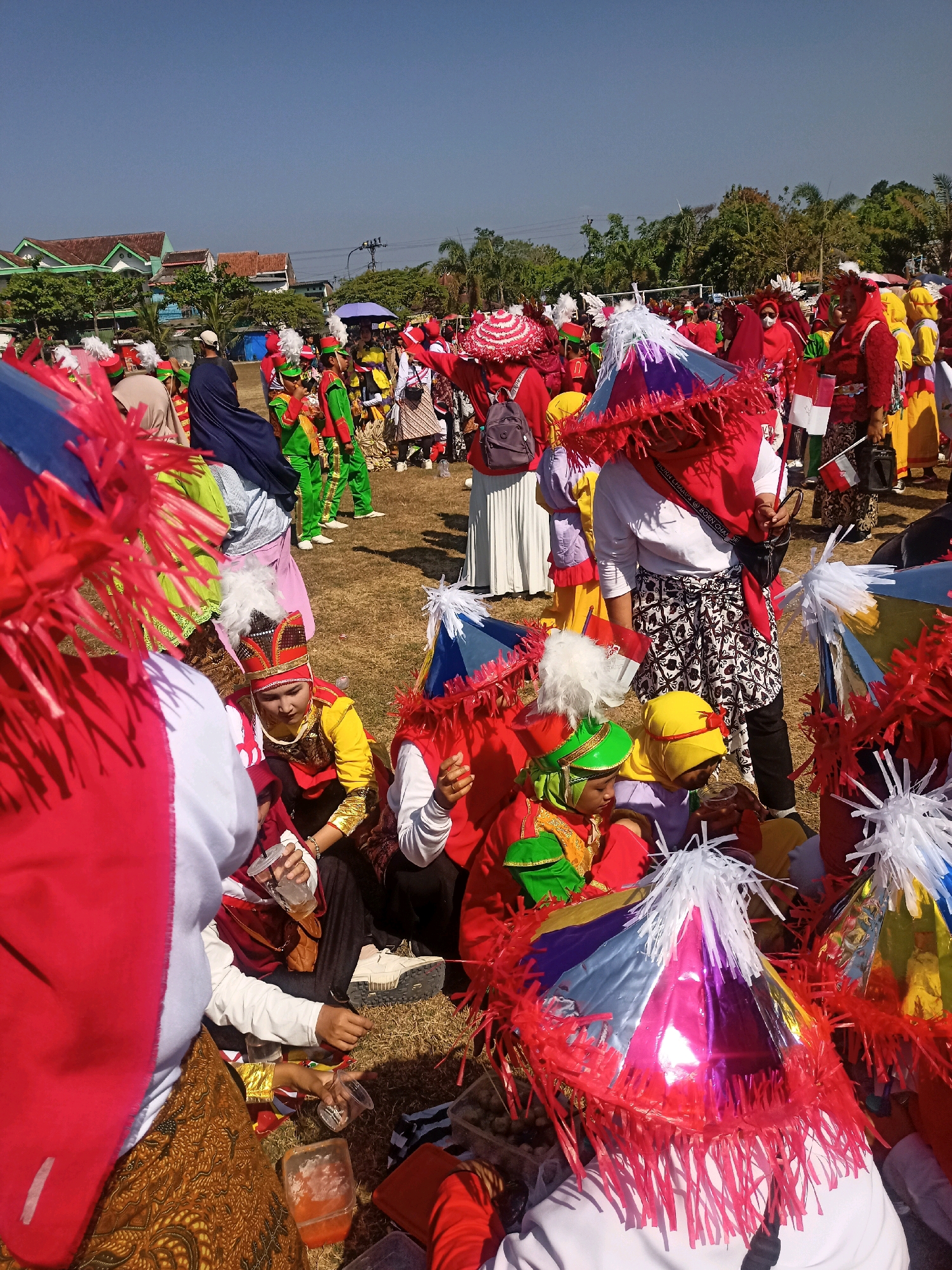 The image depicts a gathering of people in Sumberlawang, Indonesia, on a grassy field under a clear blue sky. Many individuals are dressed in colorful costumes, suggesting a festival or cultural event. Several people are wearing elaborate hats or headdresses, some adorned with white plumes. One person is wearing a yellow and red outfit with a decorative headdress. Others are in green and yellow attire. There are also people in red and white attire, some holding small flags. Decorative, cone-shaped objects with red fringe are prominent in the foreground, possibly part of the costumes or decorations.
The scene appears to be informal, with some individuals sitting on the ground. In the middle ground, there are buildings with distinctive roofs, and trees can be seen in the background. The ground is covered with dry grass and some scattered leaves. People are interacting with each other, and some are holding transparent cups. One person is holding a dark bag. The overall impression is one of a vibrant community event.