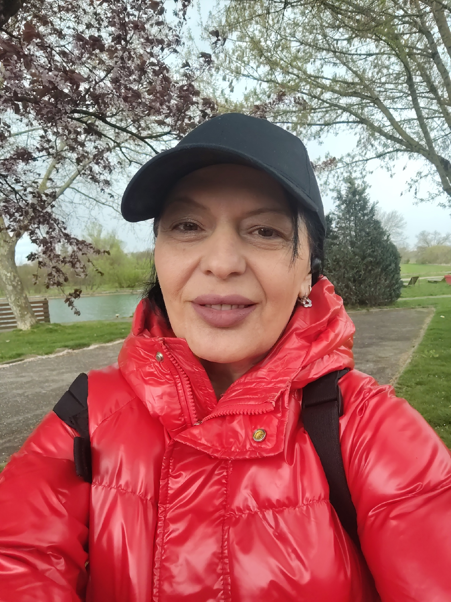 A single female subject is depicted in a close-up, selfie-style photograph taken outdoors in Souffelweyersheim, France. The subject is a middle-aged woman with dark hair, dark eyes, and light-toned skin, wearing a black baseball cap and a glossy red puffer jacket with a visible zipper and gold-colored snap buttons. A black strap, likely from a bag, is visible over her right shoulder. Small silver earrings are present. She has light pink lipstick and displays a slight smile, looking directly at the camera. The background reveals an outdoor park-like environment under an overcast sky. To the subject's left, a body of water with a light green surface is visible, partially obscured by a deciduous tree bearing reddish-brown leaves or blossoms. A wooden slatted bench is situated near the water's edge. To the subject's right, a paved path is visible, bordered by green grass. Additional trees, including a larger deciduous tree with light branches and a darker evergreen shrub, are in the mid-ground. The distant horizon shows further tree lines.