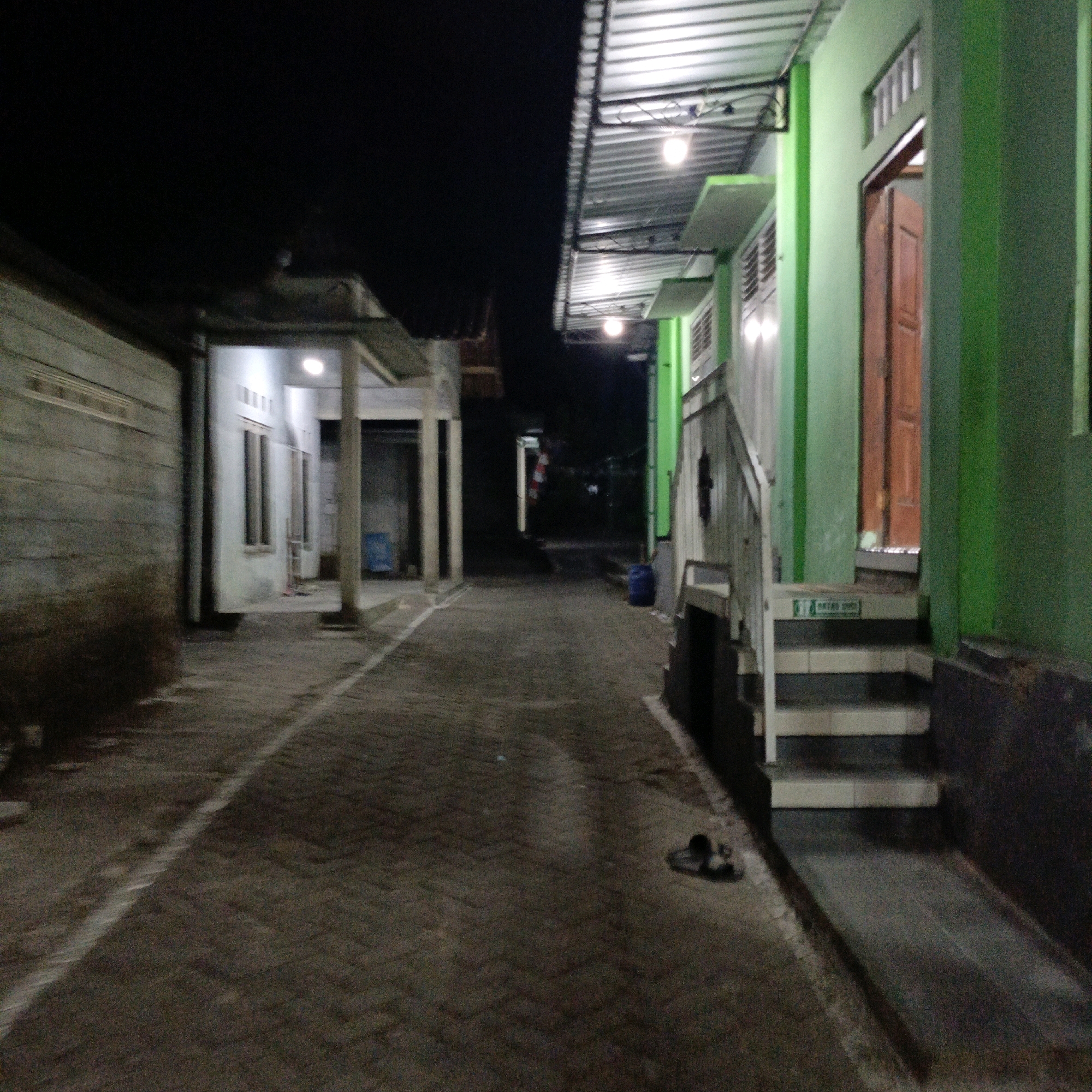 The image depicts a nighttime outdoor scene of a paved walkway between two buildings, likely in Salatiga, Indonesia. On the left, a long, low building with a concrete wall and a horizontal band of ventilation is visible. On the right, a building with a bright green facade and a covered walkway with a corrugated metal roof is prominent. The walkway is paved with interlocking bricks. Several outdoor light fixtures are visible, illuminating the area.
The right side of the image features a wooden door set within the green wall, with a small landing and a set of tiled stairs leading up to it. A sign with green text on a white background is affixed to one of the stairs. A pair of dark-colored sandals are placed on the walkway near the stairs. Further down the walkway, a blue plastic bin is visible near the green building. In the distance, another building or structure is partially obscured by darkness, with a red and white striped object visible. The overall impression is of a modest residential or commercial area at night.