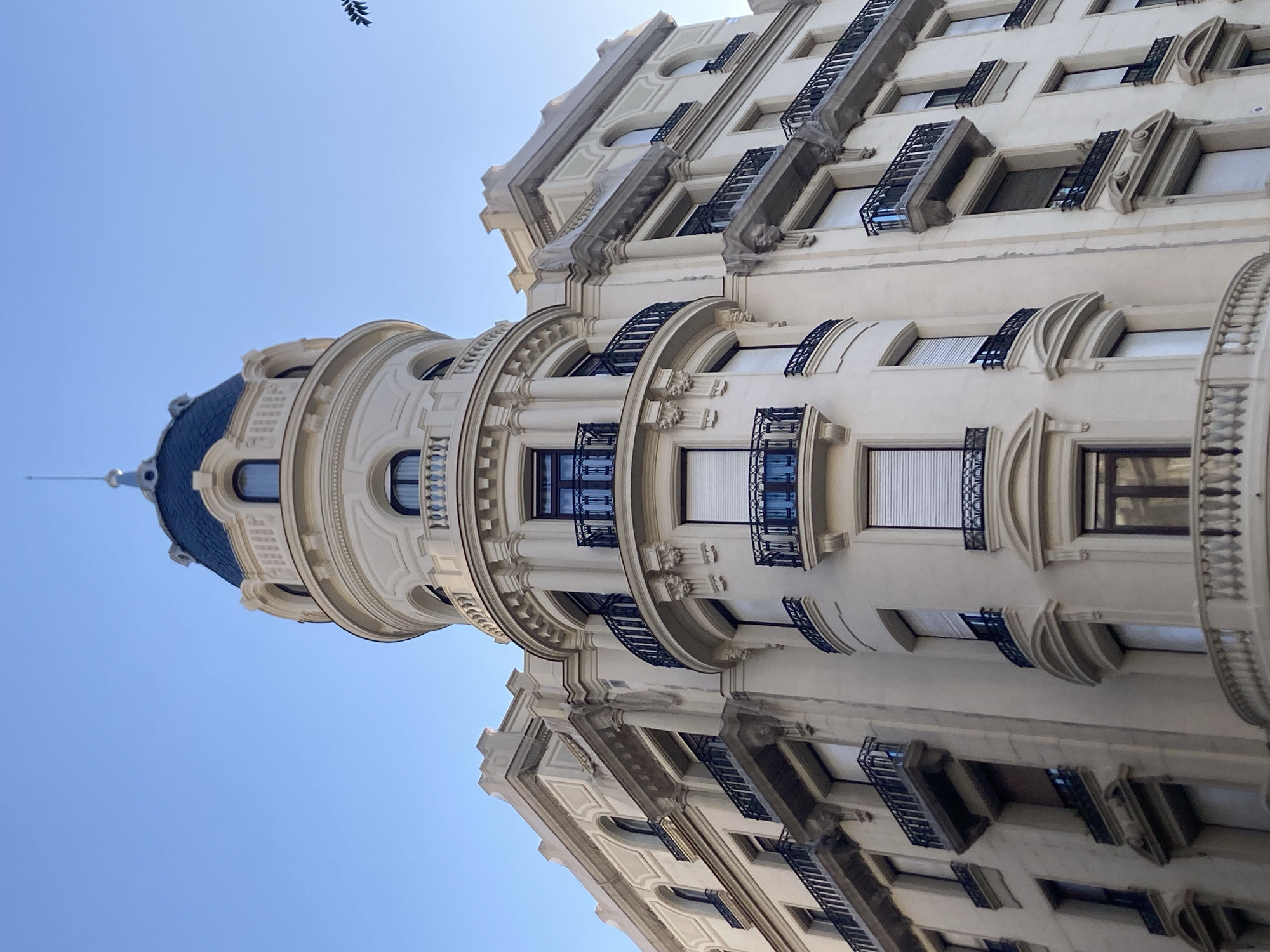 The image shows the ornate facade of a historic building, likely an apartment building, viewed from a low angle looking upwards. The architecture is grand, with cream-colored stone and detailed ornamentation. Features include rounded turrets, arched windows with black wrought-iron balconies, decorative moldings, and a blue domed roof visible on the left side of the frame. Several windows are visible, some with internal blinds drawn. The building appears to be well-maintained.
The scene is set against a clear, bright blue sky, suggesting a daytime setting, possibly late morning or early afternoon. There are no people or specific activities visible, implying the image is focused solely on the architectural details. The style of the building, with its elaborate embellishments and domed turret, evokes a sense of old-world European charm, consistent with a location like Barcelona, Spain. There is a faint, dark silhouette of a branch or leaf in the upper portion of the sky, adding a subtle natural element to the urban scene. No text is visible in the image.