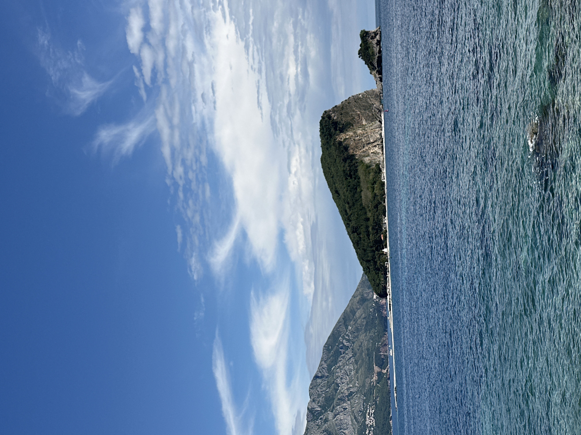 The image depicts a coastal landscape in Budva, Montenegro. The scene features a clear blue sky with scattered cirrus clouds above a mountain range. The peaks are covered with green vegetation. The sea is turquoise.