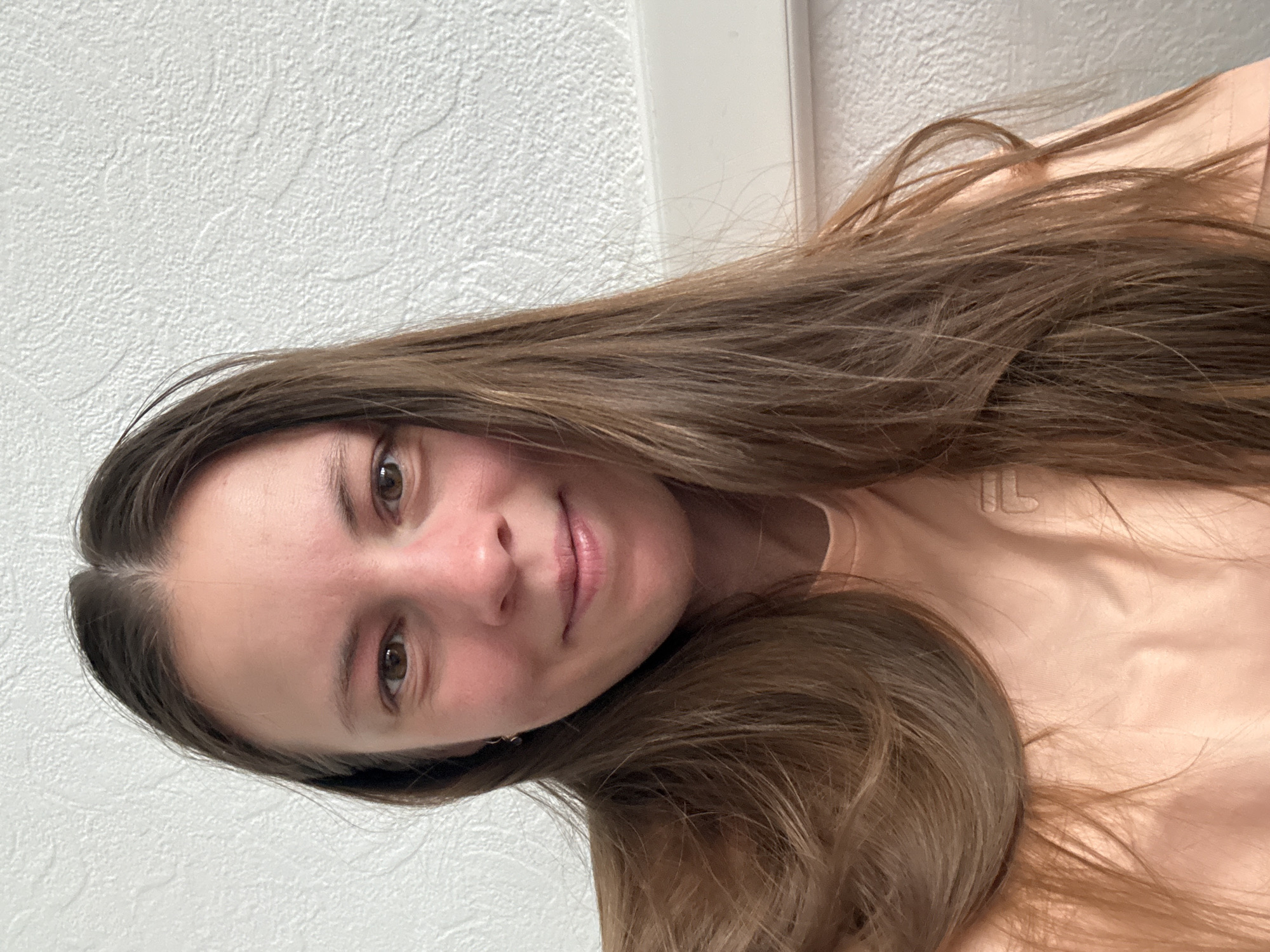 A young woman is depicted in a close-up, slightly rotated portrait shot, likely taken indoors in Chaikovsky, Russia. She has long, straight brown hair with lighter highlights, parted down the middle, which cascades over her shoulders and chest. Her hair appears thick and healthy.
She is looking directly at the camera with a gentle, slight smile. Her eyes are brown, and her skin tone is fair. She is wearing a light peach or blush-colored top, possibly a t-shirt, which has a subtle, embossed or embroidered "FILA" logo visible on the upper right side of her chest.
The background consists of a textured, light-colored wall, likely white or off-white, with a subtle stucco or popcorn ceiling-like finish. A vertical white trim or doorframe is partially visible behind her hair on the right side of the image. The lighting appears soft and even, suggesting an indoor setting without harsh direct light. The overall mood of the image is calm and informal, consistent with a casual selfie or portrait. No specific activity beyond posing for the photograph is evident. There are no strong indications of the time of day or weather beyond the indoor lighting.