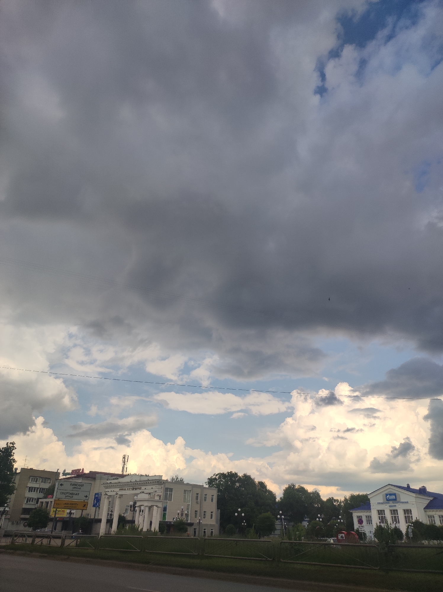 The image shows a view of the city of Chaikovsky, Russia, with buildings and trees visible under a dramatic sky filled with dark clouds and some patches of blue. On the left, a multi-story building with balconies is visible, with a sign pointing to "Kamskaya Street" and "Sportivnaya Street." Next to it is a white building with columns and a sign that reads "Kamchatka Entertainment Center." On the right, a building with a blue roof and the Gazprom logo is seen. A road and a green fence are in the foreground. Several birds are flying in the sky. The overall scene suggests an overcast day with a possibility of rain.