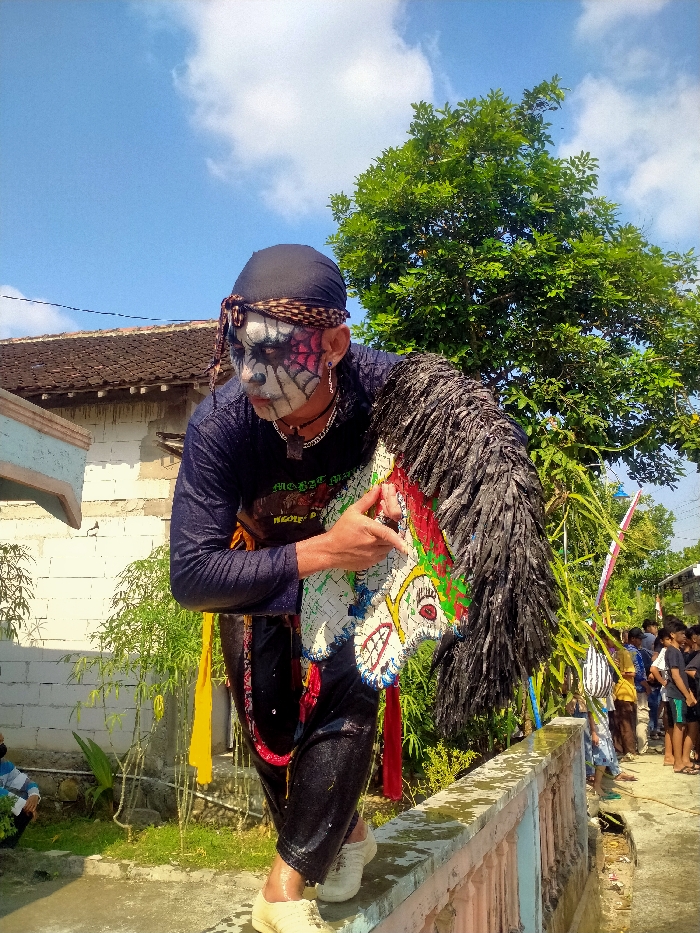 The image captures a male performer engaged in a cultural activity in Sepat, Indonesia, under a clear daylight sky. The individual is positioned centrally, leaning forward with his right foot placed on a low, wet, light-colored concrete wall. He has elaborate face paint featuring white, black, and red details, including a spiderweb pattern on his left cheek, and dark rings around his eyes. He wears a black head covering secured by a patterned brown bandana, silver hoop earrings, and a dark pendant necklace. His clothing consists of a wet, dark long-sleeved shirt and wet dark pants, with a yellow fabric sash visible around his waist and red fabric around his right leg. He holds a decorative 'kuda lumping' (flat horse) prop in his right hand, which features a white woven body with red, green, and blue accents, and a shaggy black mane. In the background, a traditional building with a dark tiled roof is visible to the left. To the right, lush green foliage and tall stalks are present, beyond which a group of people stand on a dirt path, suggesting a public event. The sky is bright blue with scattered white clouds.