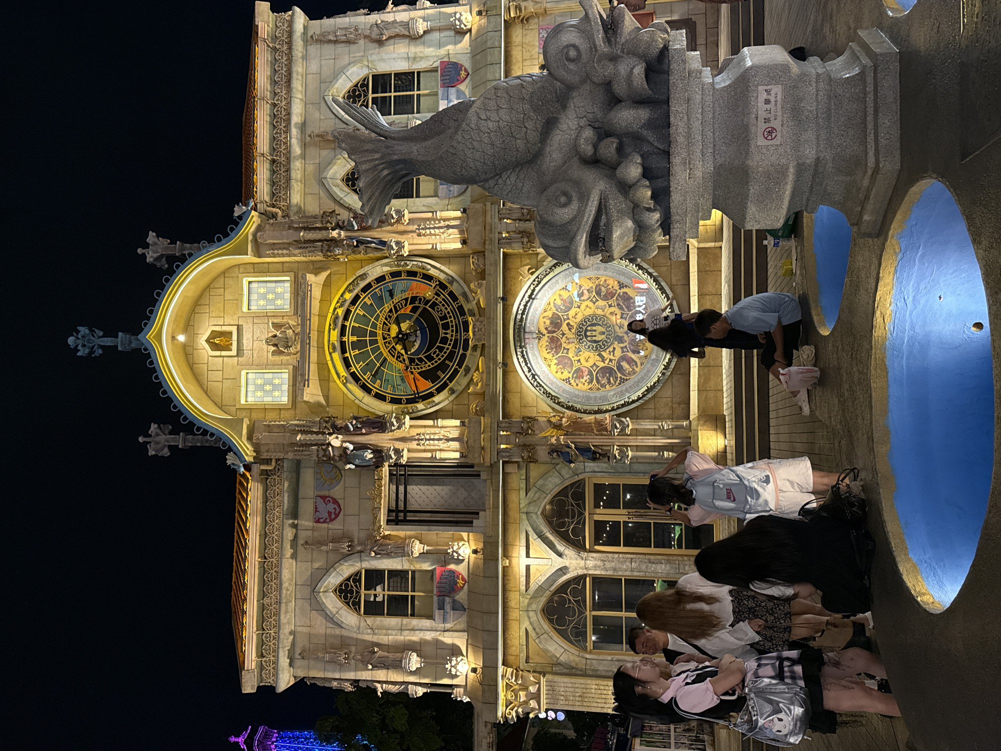 At night, a group of people stand in front of a large, ornate building that resembles a clock tower, located in the city of Nanshan District, China. The building is illuminated with warm lights, highlighting its intricate architectural details, including gothic-style windows and sculptures. A large, circular clock face is prominently displayed on the building's facade, along with a smaller circular design below it. In the foreground, a stone sculpture of a fish-like creature sits atop a pedestal, with two illuminated blue pools in front of it. Several individuals are captured in candid moments: some are looking at the building, one is taking a picture, and others are sitting or standing in conversation. The overall atmosphere is that of a public gathering space, possibly a tourist attraction, in an urban environment.