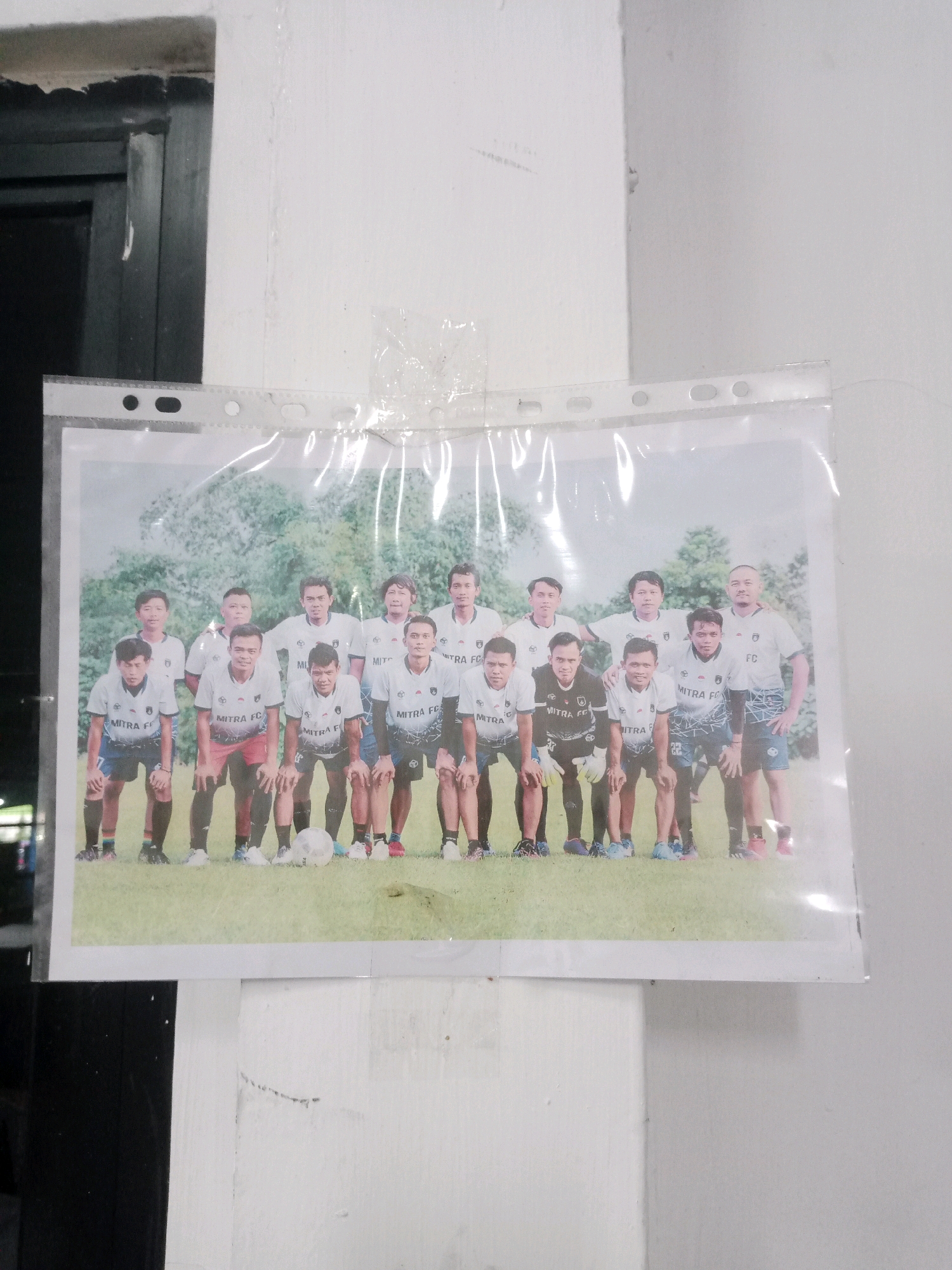 The image shows a laminated photograph of a soccer team posing for a group picture. The team is composed of 11 young men wearing white soccer jerseys with "MITRA FC" printed on them and blue shorts. Some are in the front row, crouching, while others are in the back row, standing. A soccer ball is visible on the ground in front of the crouching players. The photograph is taped to a white wall, which appears to be in an indoor or semi-outdoor setting with a dark doorway or opening to the left. The surrounding environment suggests a casual, possibly urban setting in Balaraja, Indonesia, given the context provided.