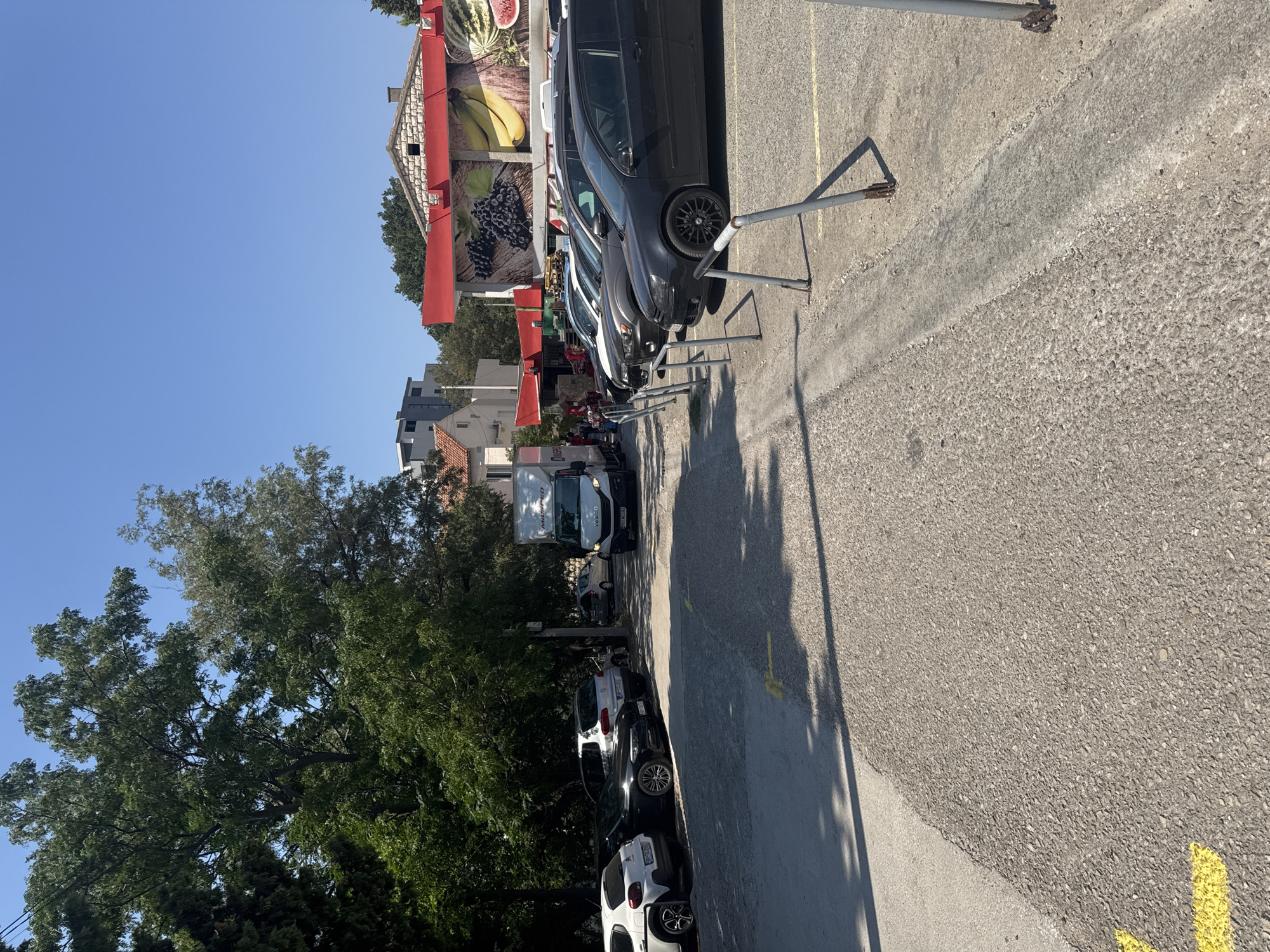 This image shows a parking lot in front of a market with a mural of fruits on its facade. A white delivery truck is parked on the right side of the frame, and several cars are parked in a row on the left. The ground is paved with asphalt, and yellow lines mark the parking spaces. Trees and buildings are visible in the background. The sky is clear and blue. The location is the market in Sutomore, Montenegro, a popular tourist destination on the Adriatic coast.