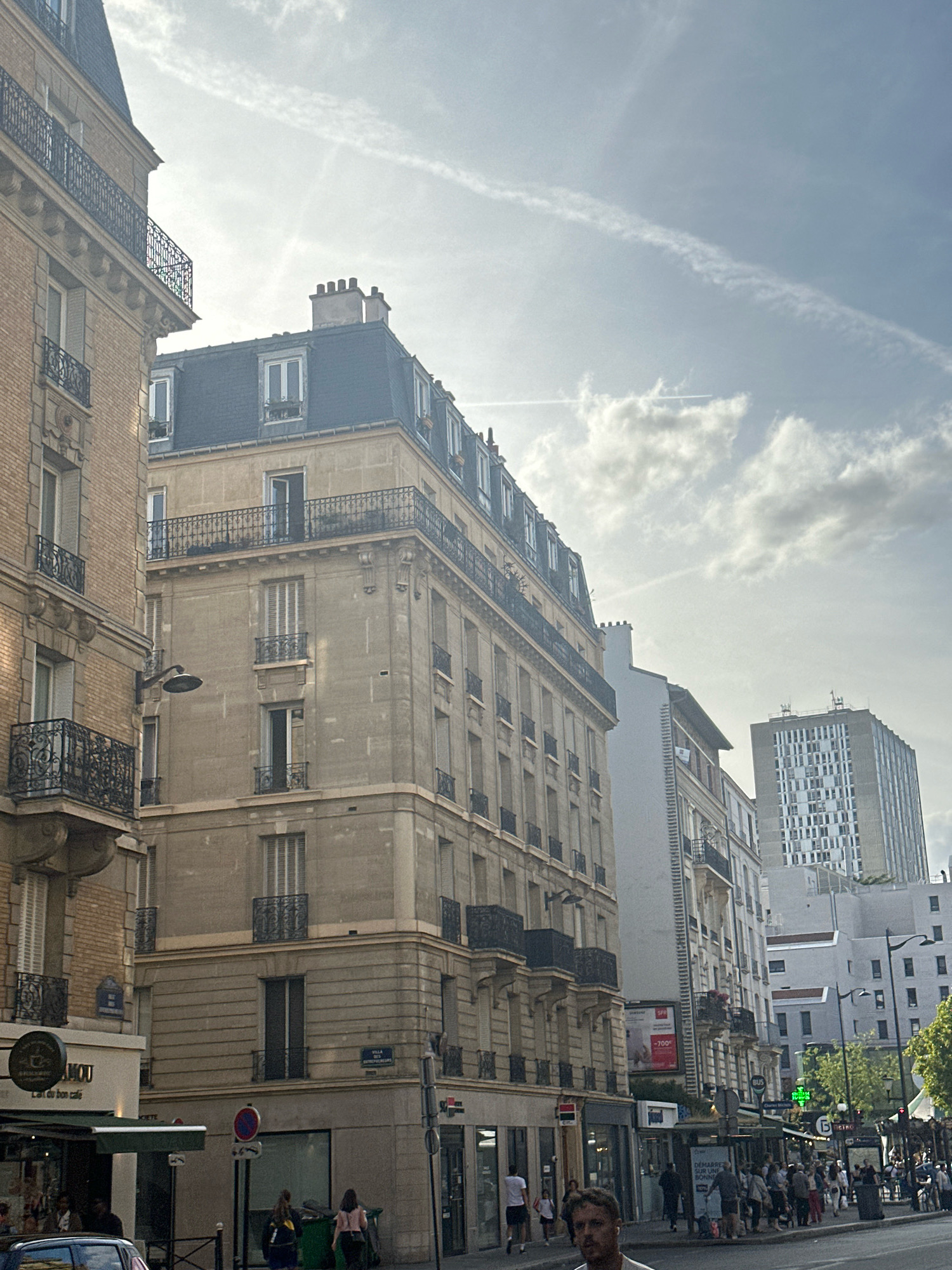 This image captures a street scene in Paris, France, likely in a commercial or residential district with a mix of older and modern architecture. The perspective is from street level, looking up at several multi-story buildings.
On the left, a tan brick building with ornate iron balconies and numerous windows dominates the foreground. Adjacent to it is a lighter-colored stone building with a mansard roof, also featuring balconies and distinct window designs. Further down the street, a taller, more modern building with a grid-like facade is visible in the background, alongside other buildings of varying heights and architectural styles.
The sky above is a mix of blue and white clouds, with visible contrails from aircraft suggesting active air traffic. A street lamp is mounted on the side of the stone building. The street itself is lined with businesses, including a cafe with an awning, and various shop fronts. Several pedestrians are visible on the sidewalk, some walking, others standing. Two individuals are seen near green trash receptacles. A car is partially visible in the lower left corner. Traffic signs and streetlights are also present, indicating a functional urban environment. The lighting suggests it is daytime, possibly late afternoon or early evening, given the angle of the light and some soft shadows. The overall impression is a bustling Parisian street with a blend of historical and contemporary elements.
