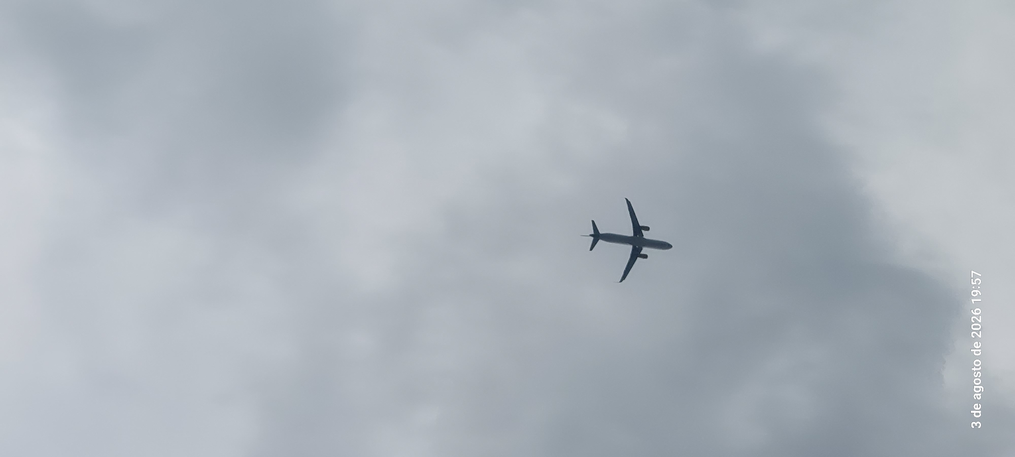 An airplane is silhouetted against a cloudy sky, viewed from below. The aircraft is flying towards the left of the frame, with its wings spread and tail clearly visible. The sky is filled with dense, grey clouds. In the bottom left corner of the image, text reads "3 de agosto de 2026 19:57", indicating the date and time. This scene could be observed anywhere, including over the city of Castelldefels, Spain, given its coastal location and proximity to airports.