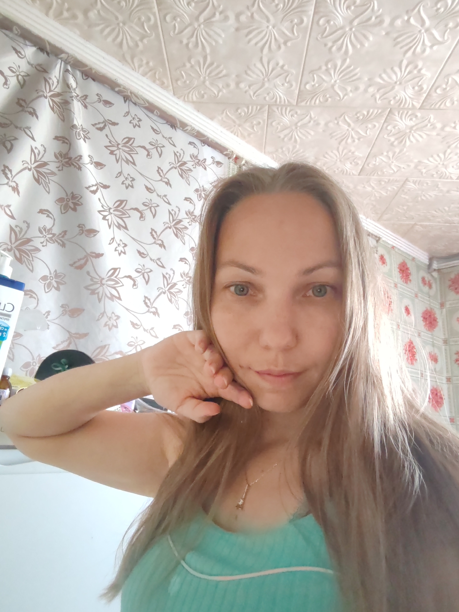 A medium shot, eye-level selfie captures a young Caucasian woman with long, light brown hair and light blue eyes. She is wearing a turquoise tank top and a delicate silver necklace. Her right arm is bent, with her hand resting gently on her cheek and chin. She is looking directly at the camera with a neutral expression. The background shows a ceiling with textured, embossed tiles and floral wallpaper in muted tones. On the left, part of a white curtain with a brown floral pattern is visible, along with a white bottle with a blue label. The overall lighting is soft, suggesting an indoor setting. The image is not specifically indicative of any location within the city of Чухлэм, Russia, or any other specific urban environment.