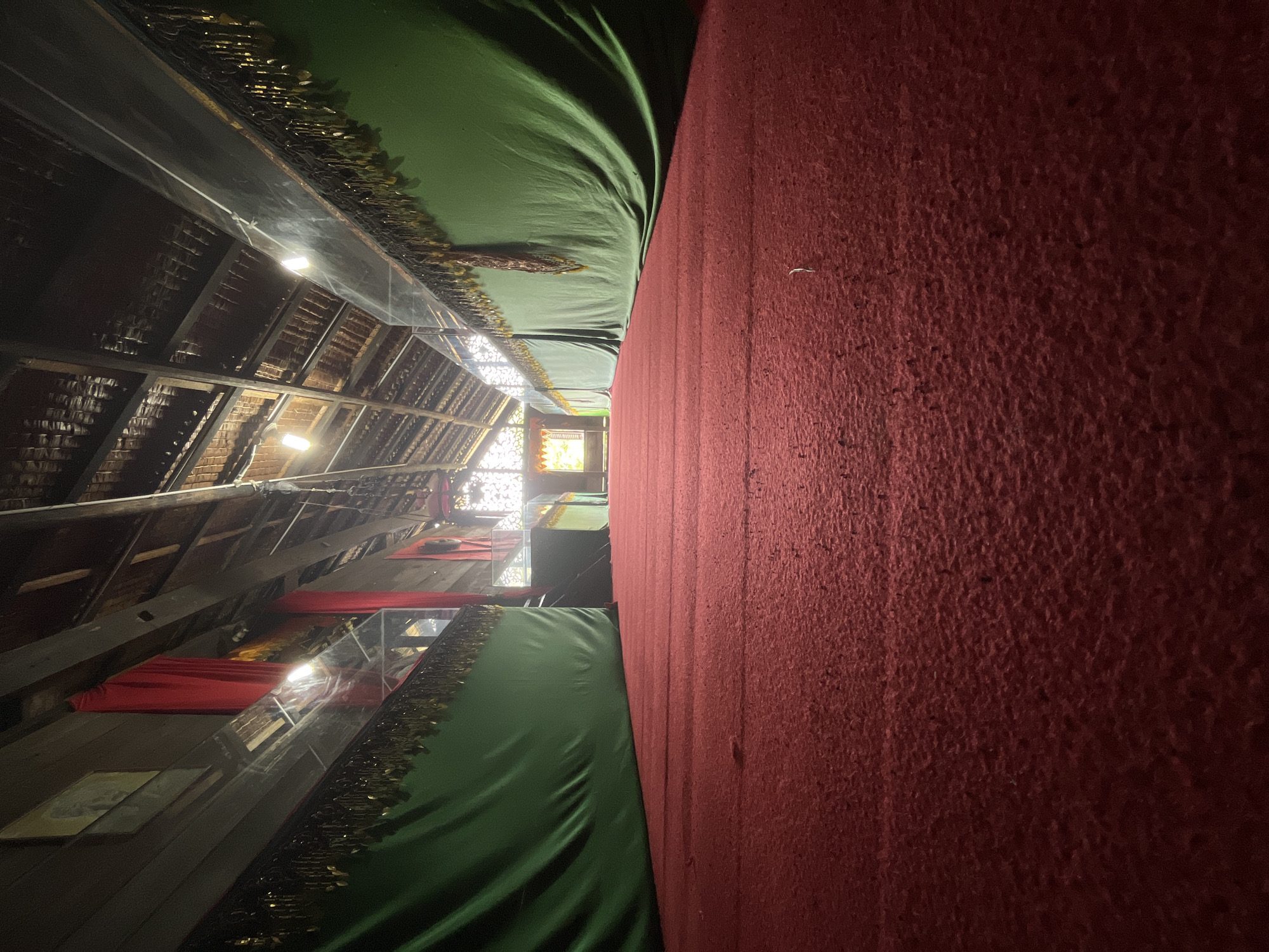 The image captures the interior of a building with a red carpet covering the floor, extending into the distance. The ceiling is constructed of exposed wooden beams and roofing material, with light fixtures visible. Along the sides of the space, green fabric drapes are hung, adorned with gold fringe. Red fabric curtains are also visible in the background. The scene appears to be within a building in Langsa, Indonesia, possibly a community hall or a place of worship, given the decorative elements. There are no people or animals present in the image, and the focus is on the architectural elements and the red carpet.