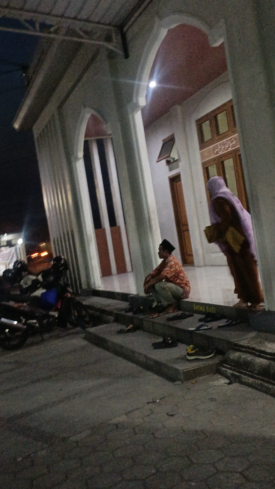 The image depicts the exterior of a building with Islamic architectural elements, possibly a mosque, at night. A man wearing a black cap and a patterned shirt is crouched on a step. To his right, a person in a purple hijab and brown robe stands, holding an object. Several pairs of sandals and shoes are placed on the steps and ground. In the background, motorcycles are parked, and vehicle lights suggest a road nearby. A sign with Indonesian text, "BATAS SUCI" (meaning "Sacred Boundary"), is visible on a step. The overall environment appears to be an outdoor area adjacent to the building. The location is identified as Ngaru Aru, Indonesia.