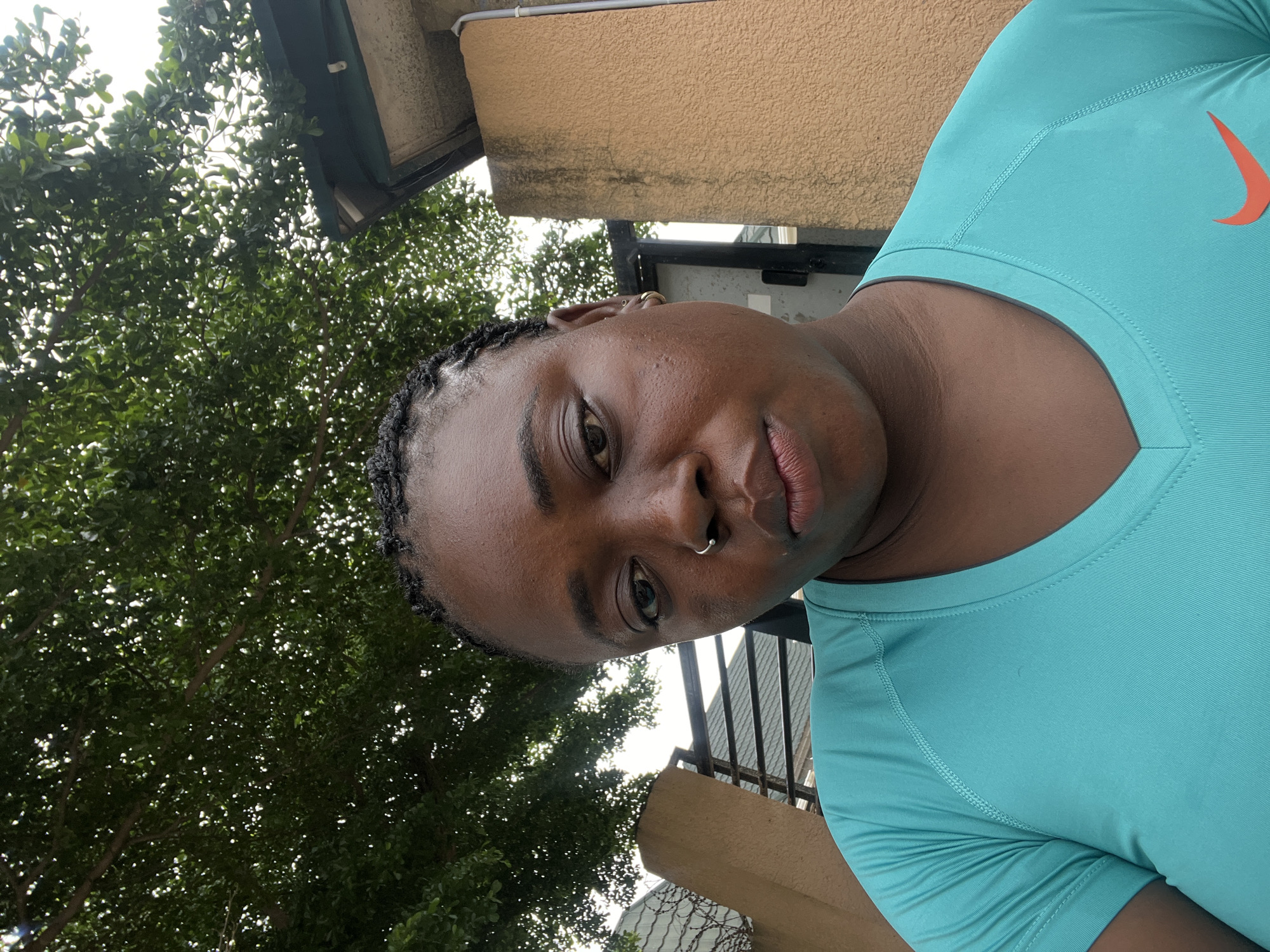 A medium shot, taken from below eye level, shows a Black woman with braided hair and a nose ring. She is wearing a turquoise v-neck t-shirt with a Nike logo on the right side of her chest. Her skin is dark, and she has a stern expression on her face. The background shows a tree with green leaves and a light-colored building. The image appears to have been taken outdoors in a residential area, possibly in Abuja, Nigeria.
