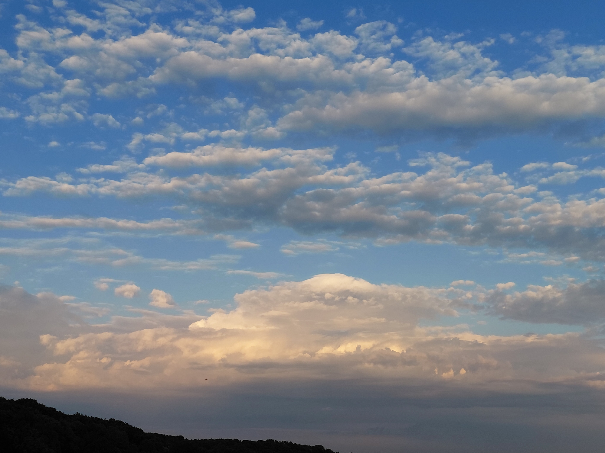 The image depicts a wide, horizontal view of a sky filled with various types of clouds. The sky is a vibrant blue, with the clouds exhibiting a range of white, grey, and golden hues, suggesting the time of day is either sunrise or sunset. A dark, silhouetted treeline or hillside occupies the very bottom of the frame, obscuring the ground level. The clouds are layered, with some appearing scattered and puffy, while others form more defined bands or masses. One particularly large, luminous cloud formation is visible in the middle ground, illuminated by warm light. The overall atmosphere is serene and natural, with no people or man-made structures readily apparent in the sky itself. The location is identified as Verkhnyaya Podstepnovka, Russia.