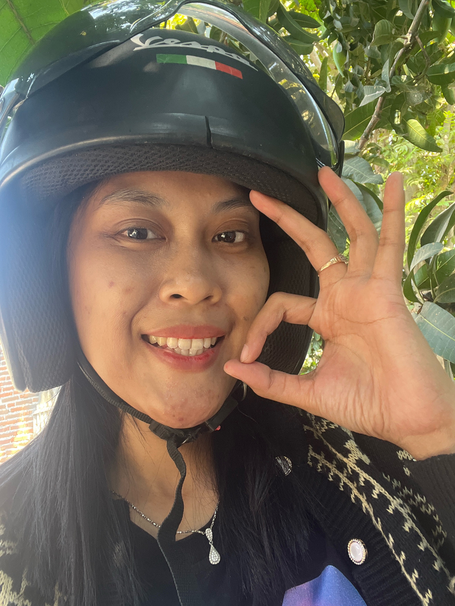 An overhead, close-up, selfie-style shot shows a woman from the chest up. She is wearing a black helmet with a white stripe that includes the Italian flag colors. The woman has dark hair and is smiling with her mouth open, showing her teeth. Her right hand is in the frame, with her thumb and index finger forming a circle, with her other fingers extended. She is wearing a gold ring on her right ring finger. She is wearing a black sweater with a white, abstract pattern on the shoulders and chest, and a silver necklace with a teardrop-shaped pendant. The background is blurred green foliage, suggesting an outdoor environment.