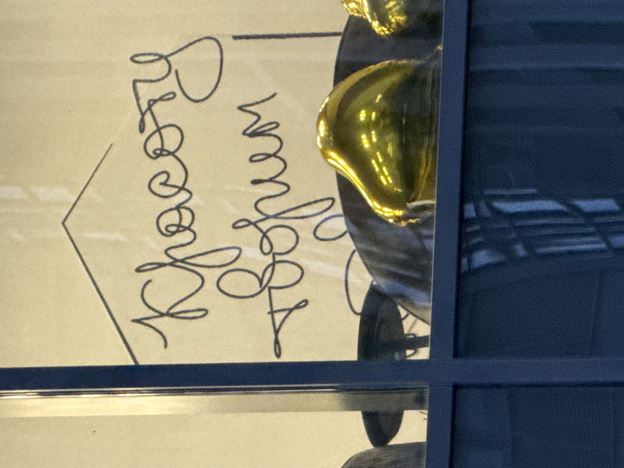 The image shows a handwritten message on a light-colored surface, possibly a card or paper, with a dark, stylized drawing next to it. The text is in a flowing, cursive script. The first word appears to be "Peace" or a similar word starting with "P" followed by an "e" and "a". The second word is less clear, but might be "on" or "in". The drawing is a stylized house or arrow pointing upwards and to the left. To the right of the paper, there is a shiny, golden object, possibly a decorative element or part of a sculpture, reflecting light. The background is dark blue, suggesting a window or frame, and there are reflections of interior lighting or a room on the shiny object. The setting appears to be indoors, likely a display or a shop window, given the polished surfaces and reflective elements. The time of day cannot be determined from the image. There are no people visible, but the presence of a decorative item and handwritten message suggests a context of gifting, celebration, or artistic display. The overall impression is one of elegance and artistry.