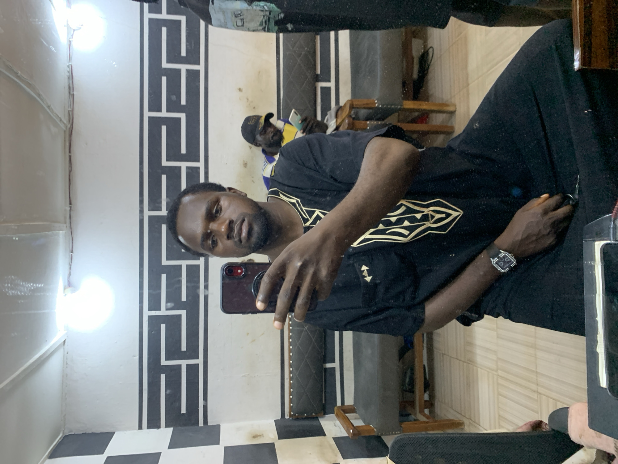 In a dimly lit room, likely a barbershop or similar establishment in Jos, Nigeria, a man is captured taking a mirror selfie. He is wearing a black traditional African garment with gold embroidery around the neckline. He holds a dark-colored smartphone in his right hand, facing the mirror to capture his reflection. A watch with a dark band and a square face is visible on his left wrist.
The background reveals a tiled floor and a wall adorned with a distinctive black and white geometric pattern above a dark upholstered bench. To the left, a black and white checkered tile pattern is partially visible. Further in the background, another man wearing a black baseball cap and a yellow and blue striped shirt is seated, looking at his own phone. The room appears to be a waiting area, with additional seating and furniture visible in the reflection. The overall lighting suggests artificial illumination from above, with a bright light source casting reflections on the ceiling. The scene is orderly and functional, typical of a service-oriented establishment.