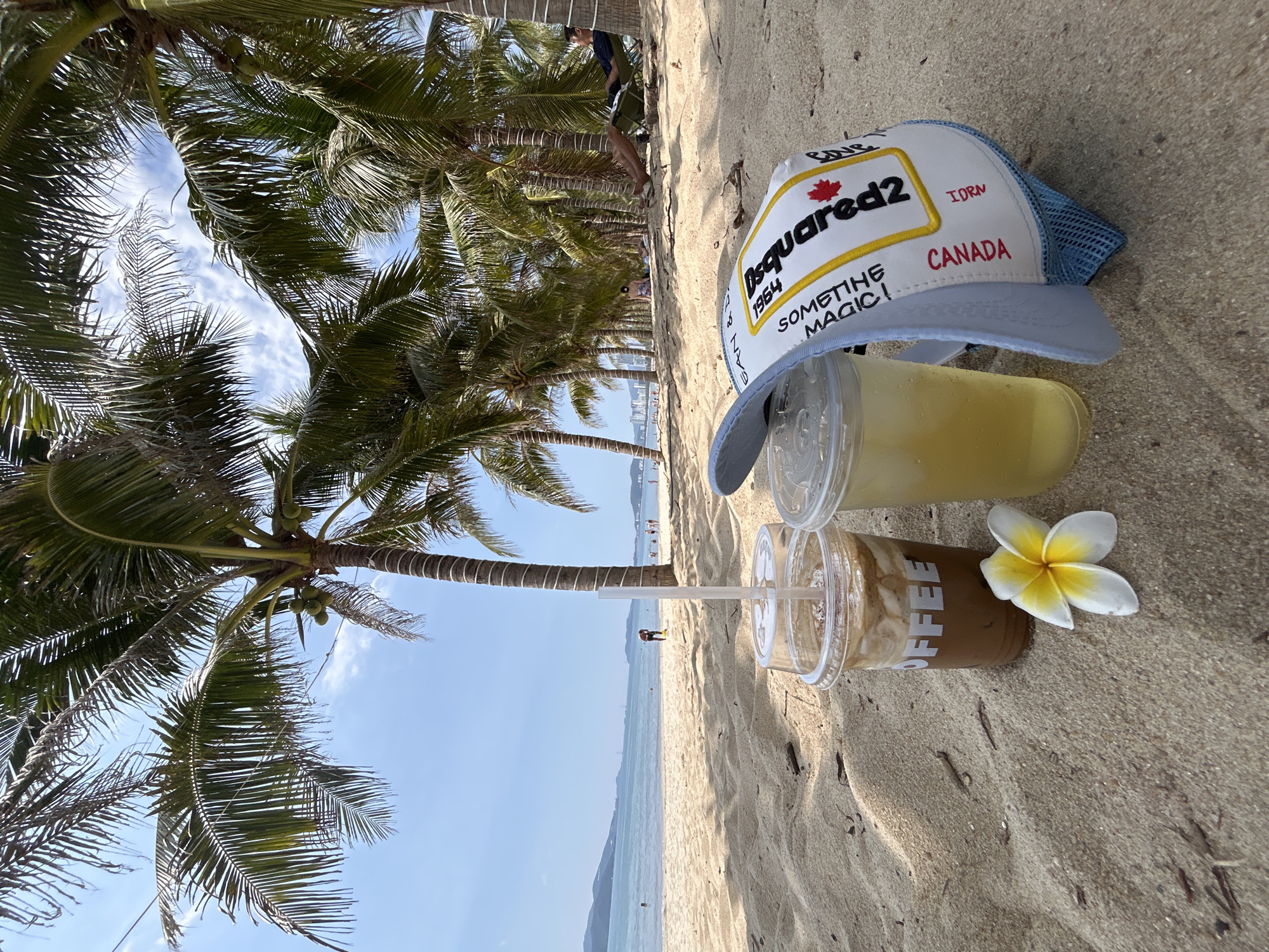 The image shows a beach scene in Nha Trang, Vietnam. A sandy beach is present with palm trees lining the shore. A clear blue sky and calm sea are visible in the background. Two plastic cups, one containing a yellow liquid and the other a brown liquid labeled "COFFEE," are resting on the sand, alongside a white and yellow plumeria flower and a white baseball cap with "Dsquared2 CANADA" printed on it. People can be seen in the distance along the shoreline.