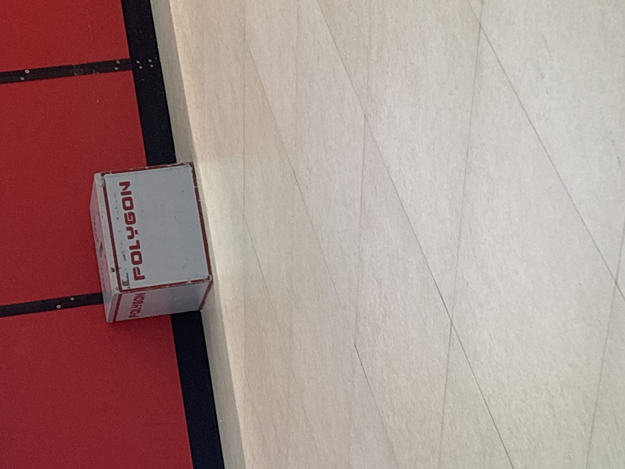 The image captures an overhead view of an indoor floor area, featuring a small white rectangular box placed on a boundary between two distinct surfaces. On the left, a textured red surface, possibly a mat or court, is visible. This red area is bordered by a thick black line or strip, which has small, light-colored circular markings, possibly rivets or fasteners, embedded along its length.
To the right of the black line is a lighter-colored surface, appearing to be off-white or light grey, with subtle diagonal lines etched or patterned across it. This could indicate a floor material such as wood, laminate, or large tiles. The white box rests centrally on the black boundary line, with one side facing the camera.
The word "POLYGON" is clearly visible in red, capital, sans-serif letters on the side of the box. This text is oriented vertically. A second instance of "POLYGON" is visible in a smaller font size on the lower edge of the same side of the box, oriented horizontally. The box shows some signs of use or minor scuffs. No human activity or specific event is taking place. The setting appears to be an interior space, possibly a sports facility or a large open room. The provided context specifies the location as Yerevan, Armenia.