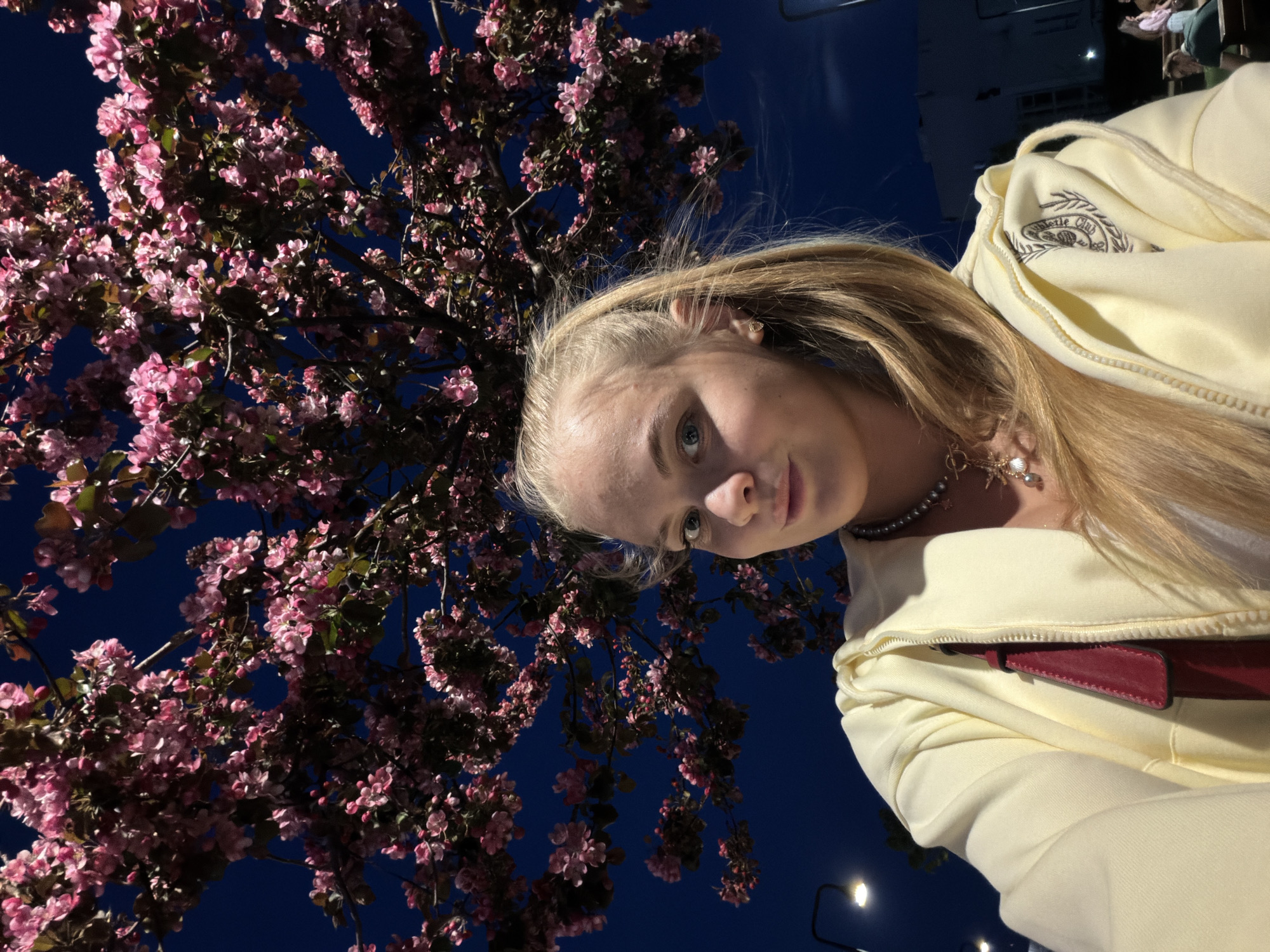 A young blonde woman stands beneath a blossoming tree at night. The tree is in full bloom with pink flowers. She wears a pale yellow hoodie with a burgundy strap. She also wears a pearl necklace. The image is likely taken in Izhevsk, Russia, based on visual landmarks. The lighting indicates it is nighttime. The sky is dark blue. There are street lamps visible.