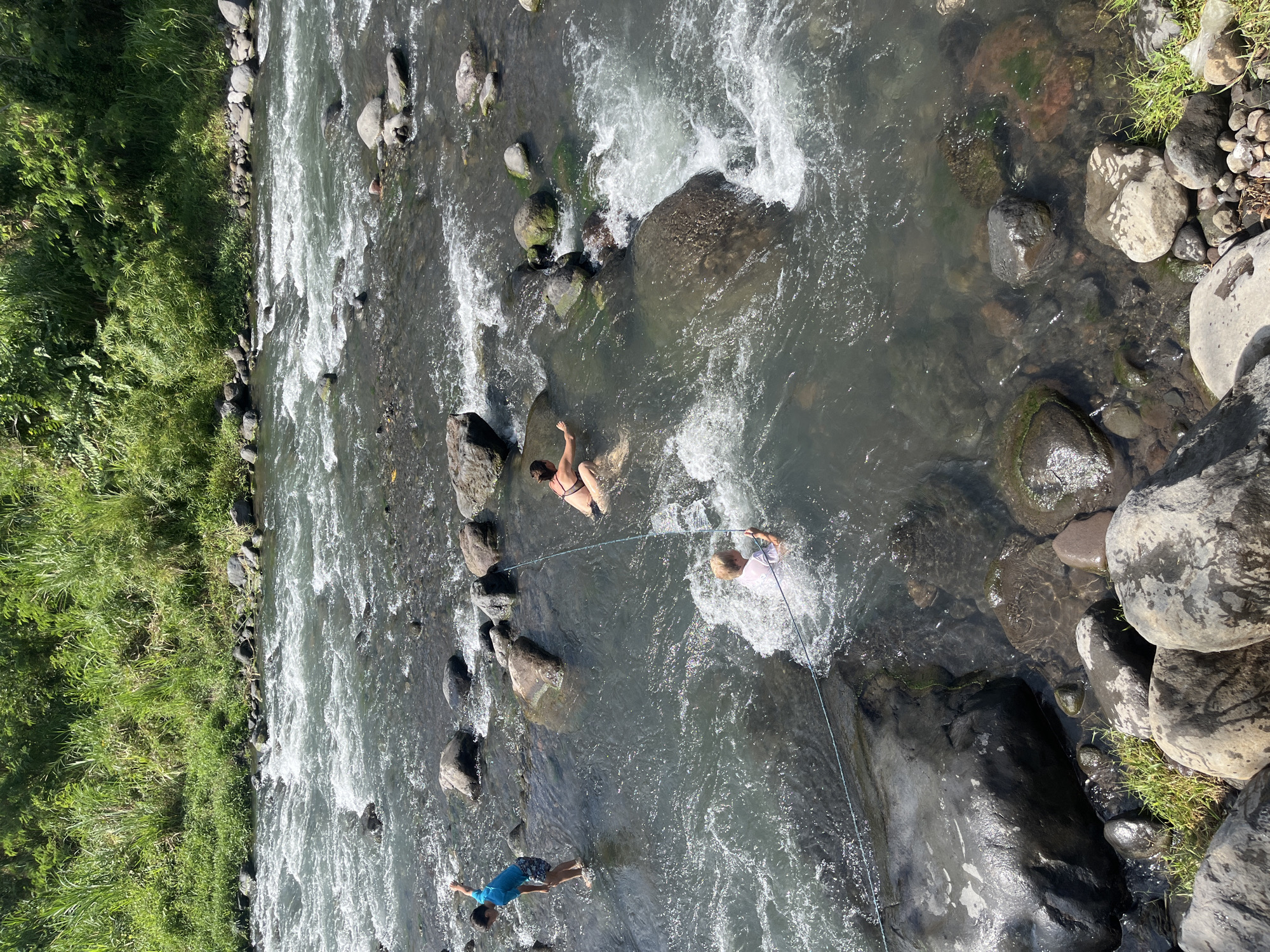 The image depicts a group of people interacting with a flowing river, likely in the Klungkung region of Indonesia. The riverbed is comprised of numerous rocks of varying sizes, with some submerged and others exposed. Lush green vegetation lines the riverbank in the background, suggesting a natural environment.
In the foreground and mid-ground, three individuals are visible in the water. A young person wearing a blue t-shirt and patterned shorts stands in the river on the left, with their right arm raised. To the right of this individual, a person in a dark bikini is partially submerged, facing away from the camera. Further to the right and slightly in front, another person in a white t-shirt is also partially submerged, with water splashing around them, and they are holding a rope. A blue rope appears to extend across the water, connecting to this individual. The water is clear enough to see the rocks beneath the surface in some areas, and there are visible ripples and currents indicating the flow of the river. The sunlight suggests a daytime scene.