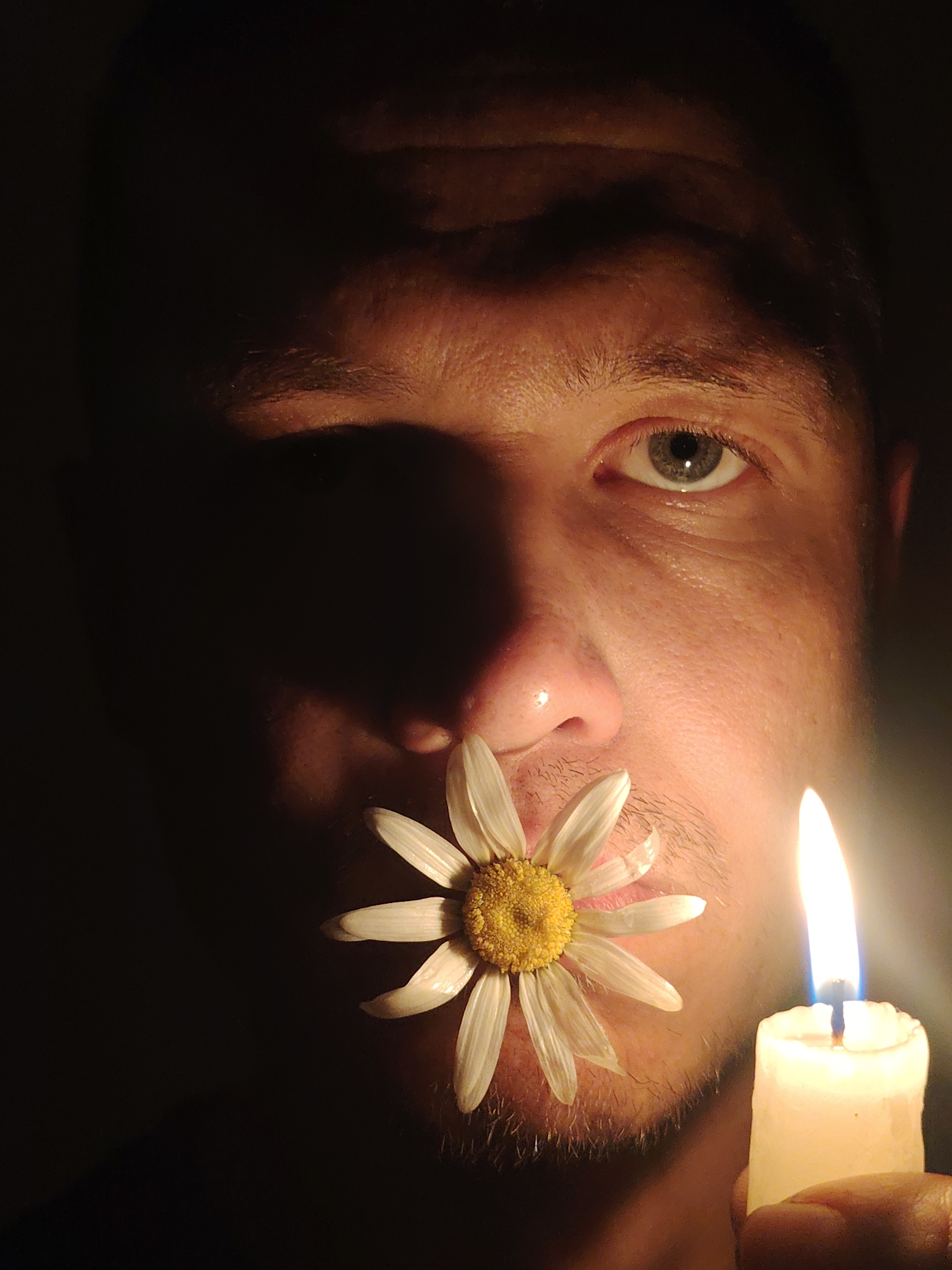 A close-up shot captures a person's face illuminated by a lit candle. A white daisy is held between the person's lips, with its petals fanning outwards. The person's right eye, which is green, is visible and looking forward. The left side of the face is in shadow, obscuring most of the features. The background is dark, with the faint glow of the candle and its flame being the primary light source. The scene appears to be indoors, given the close proximity and controlled lighting. There is no indication of the location being in Chaikovsky, Russia, as there are no recognizable landmarks or contextual clues.