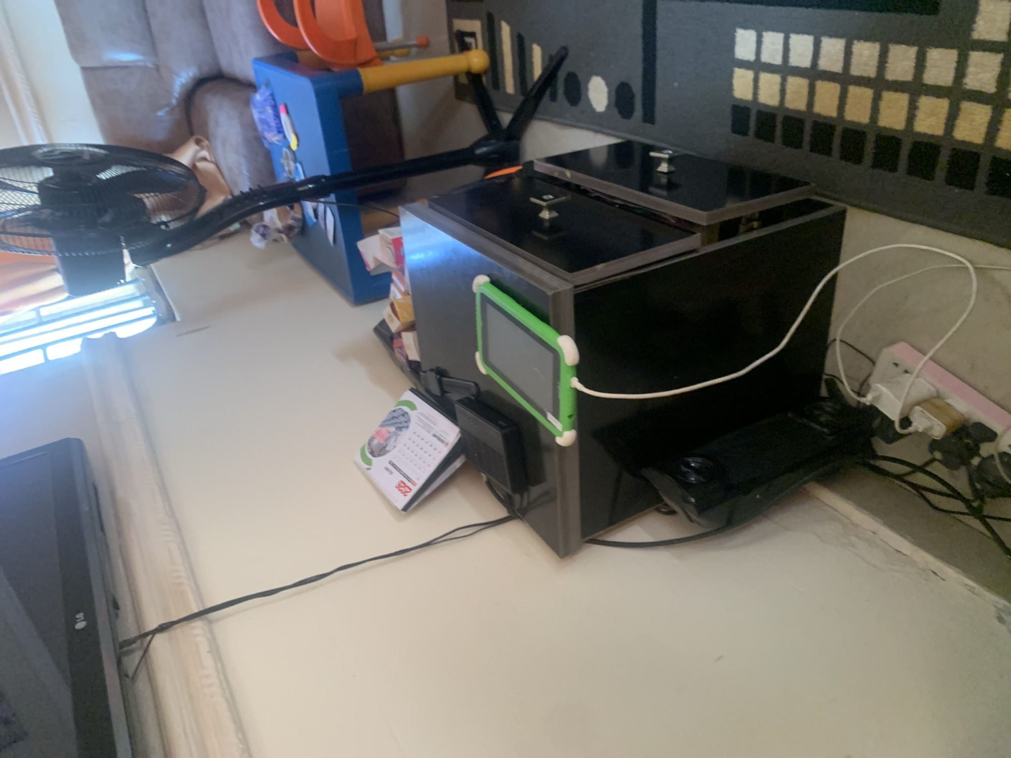 The image depicts an indoor scene, likely a living room, in the city of Gura Top, Nigeria. A television is mounted on the wall, with a black cable extending from it downwards. Below the television, on a dark cabinet, a tablet with a green case is charging via a white cable. Next to the tablet, a black electronic device and a small calendar are visible. In the background, a standing fan is positioned near a blue table with some items on it, including what appears to be a child's chair. A couch is also partially visible in the background. The floor is covered by a dark rug with a checkered pattern. Electrical cords and a power strip are present near the bottom left of the image, connected to the various devices. The overall impression is of a functional, lived-in space with electronics and furniture typical of a home environment.