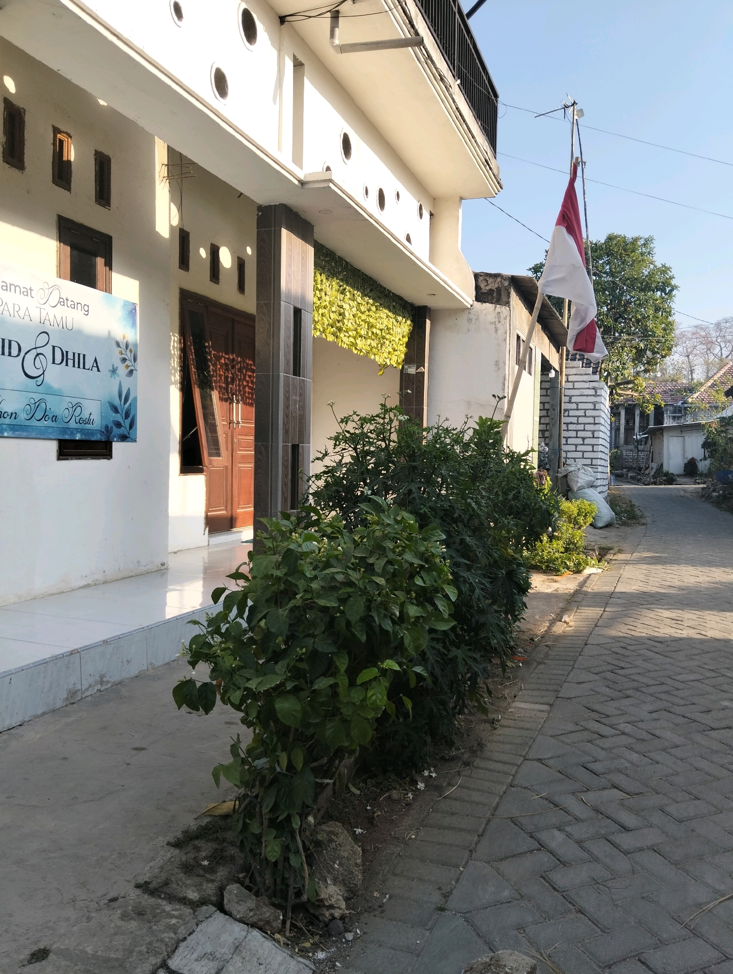 The image captures a sunlit street in the city of Drajat, Indonesia. In the foreground, a paved walkway lined with lush green bushes leads away from a white building. The building has a modern design with dark brown doors and windows, and a balcony on the upper level. A banner with text in an Indonesian language is displayed on the building's exterior. To the right of the building, an Indonesian flag is prominently displayed on a makeshift flagpole. The street continues into the distance, bordered by more buildings and trees. The overall atmosphere is one of a quiet, residential neighborhood bathed in the warm glow of daylight.