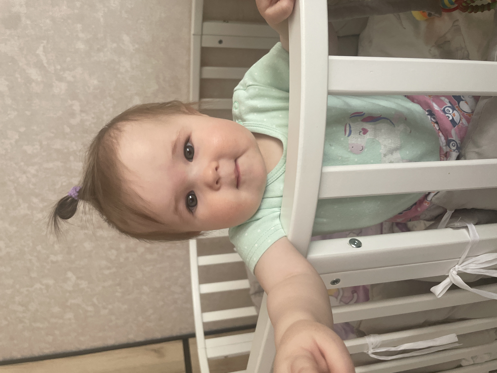 A young baby, appearing to be an infant, is shown standing in a white crib, holding onto the top rail and looking directly at the camera. The baby has light brown hair, which is styled into a small ponytail secured with a purple elastic band, and dark, expressive eyes. A soft smile plays on the baby's face, conveying a happy and curious demeanor.
The baby is dressed in a light mint green onesie patterned with small white polka dots. On the front of the onesie is a graphic of a white unicorn with a pink mane and tail. Inside the crib, patterned bedding is visible; one sheet clearly features a unicorn design, and another has a pattern of pink and blue penguins. Hints of soft toys or rattles are discernible in the background within the crib.
The white crib has horizontal bars. The background wall is covered with a light-colored, textured wallpaper. The image is captured from a slightly low angle, looking upwards at the baby, with one of the baby's arms extended outward from the crib. The scene takes place indoors, likely in a nursery or bedroom, under well-lit conditions. There is no visible text in the image. The implied location is a home in Chaikovsky, Russia.