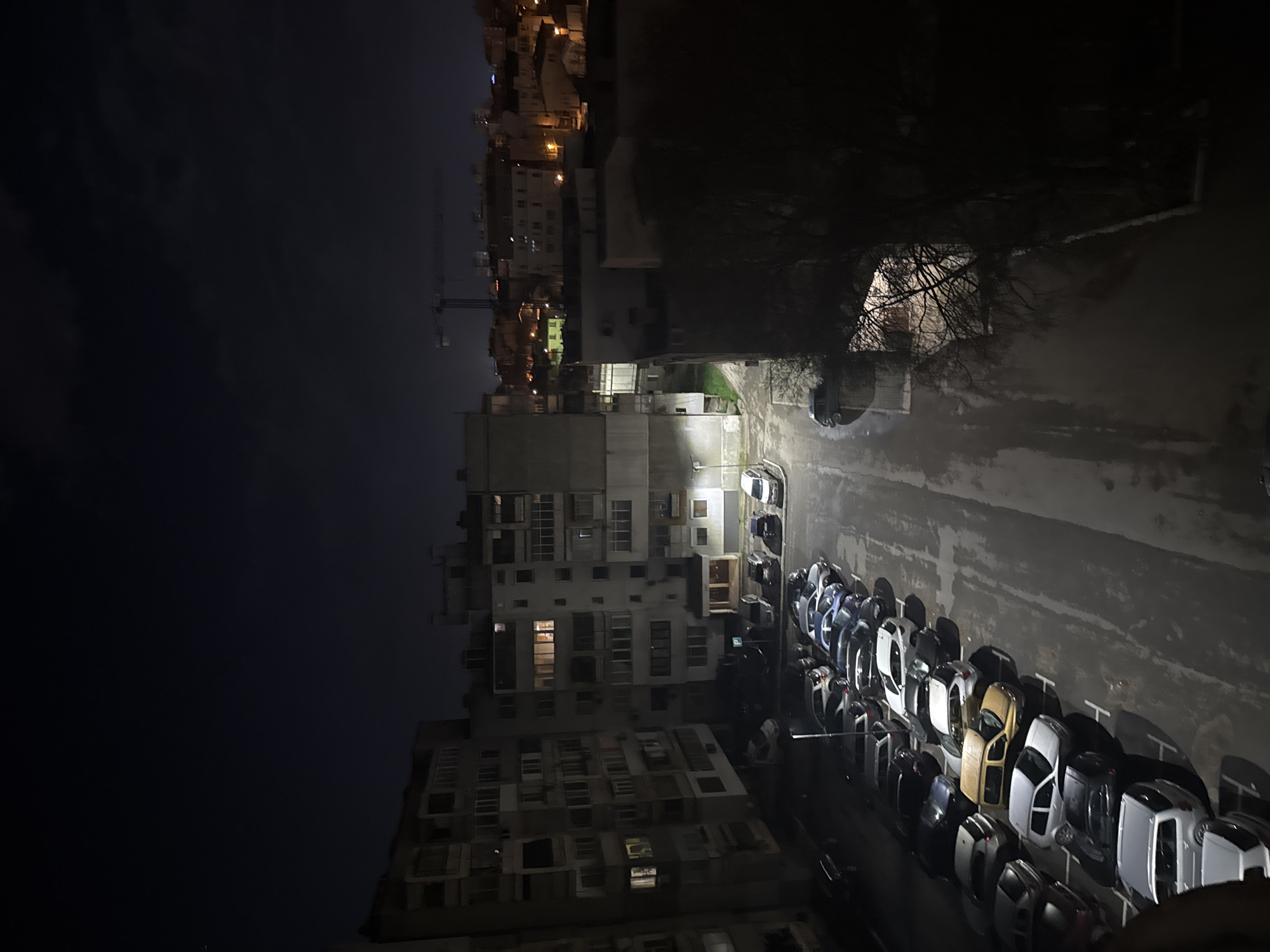 An elevated view captures a residential area in Varna, Bulgaria at night. The sky is dark and overcast. In the distance, city lights illuminate several buildings. A construction crane is visible. Closer to the viewer, a tall apartment building is centrally placed with some illuminated windows. Below, a parking lot contains numerous parked cars arranged neatly. A tree is to the right of the building. The road beside the parking lot is wet, reflecting the light.