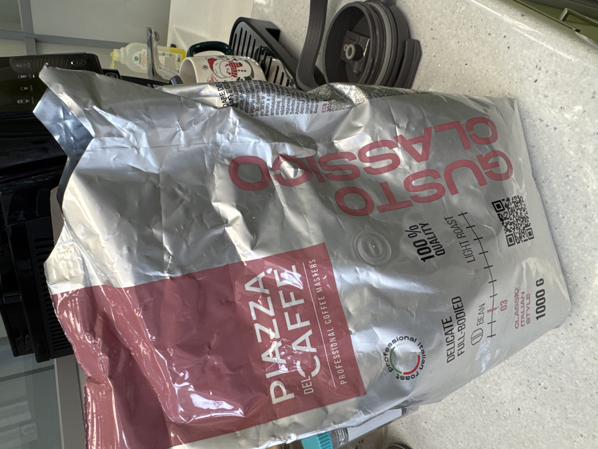 The image displays a large, silver and maroon bag of coffee beans, "Piazza Del Caffe Gusto Classico," resting on a light-colored, speckled countertop. The bag is crumpled, suggesting it is in use.
Visible text on the coffee bag includes:
* "PIAZZA DEL CAFFE"
* "PROFESSIONAL COFFEE MASTERS"
* "GUSTO CLASSICO"
* "DELICATE FULL-BODIED"
* "100% QUALITY"
* "LIGHT ROAST" (indicated on a scale)
* "BEAN" (with an icon)
* "03" (indicating roast intensity on a scale)
* "CLASSIC ITALIAN STYLE"
* "1000 G"
* A circular logo reading "professional italian roast."
* Care instructions and product information in Romanian, including "cafea", "Moldova", "Basarab", and reference to "Sverdlov.md."
* Text in Georgian characters, including "პიაც" (Piazza).
* A QR code is also visible on the bag.
In the background, behind and to the right of the coffee bag, several items are visible on the countertop. These include a black coffee machine or kitchen appliance, a white mug with a red Santa Claus design, a yellow bottle, and a grey plastic lid or component, possibly part of a coffee grinder or another kitchen item. The scene suggests a domestic kitchen setting, possibly in Moscow, Russia, where the product, with its multi-lingual packaging, is being used. No specific activity is taking place; the items are stationary. The lighting appears to be indoor ambient light.