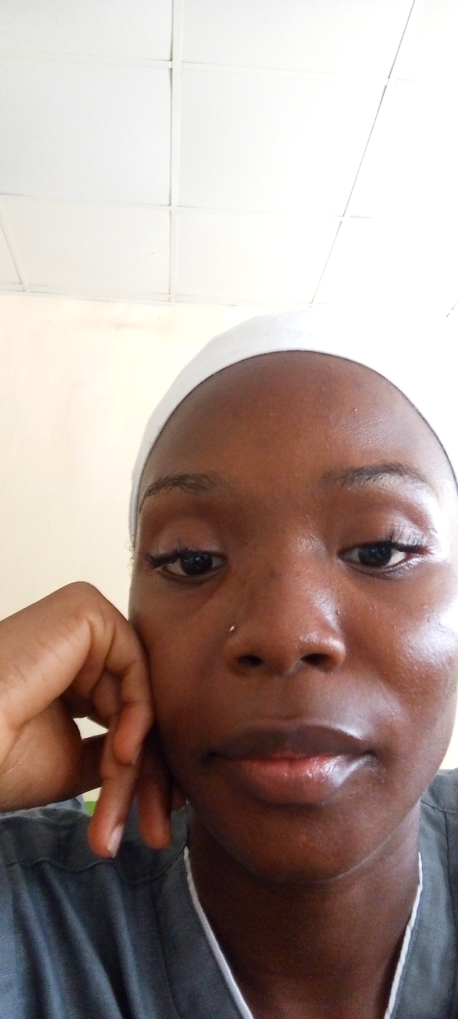 A close-up portrait features a person with dark skin, looking directly or slightly downwards towards the camera. They wear a white head covering, possibly a hijab or scarf, and a grey top with a white trim along the V-neckline. A small, silver nose piercing adorns their right nostril. Their left hand rests gently against their left cheek and jaw, with fingers visible, suggesting a contemplative or relaxed pose. Dark eyes are visible, enhanced by what appears to be subtle eye makeup. Their lips are closed, exhibiting a natural sheen.
The background consists of a plain, light-colored wall (cream or light beige) in the upper left, transitioning to a bright white suspended ceiling with a grid pattern, typical of acoustic tiles, overhead. The lighting is soft and even, indicating an indoor environment. The setting appears to be an institutional or public building rather than a private residence, inferred from the ceiling type. The person's expression is neutral, conveying a sense of calm or pensiveness. No visible text is present in the image.