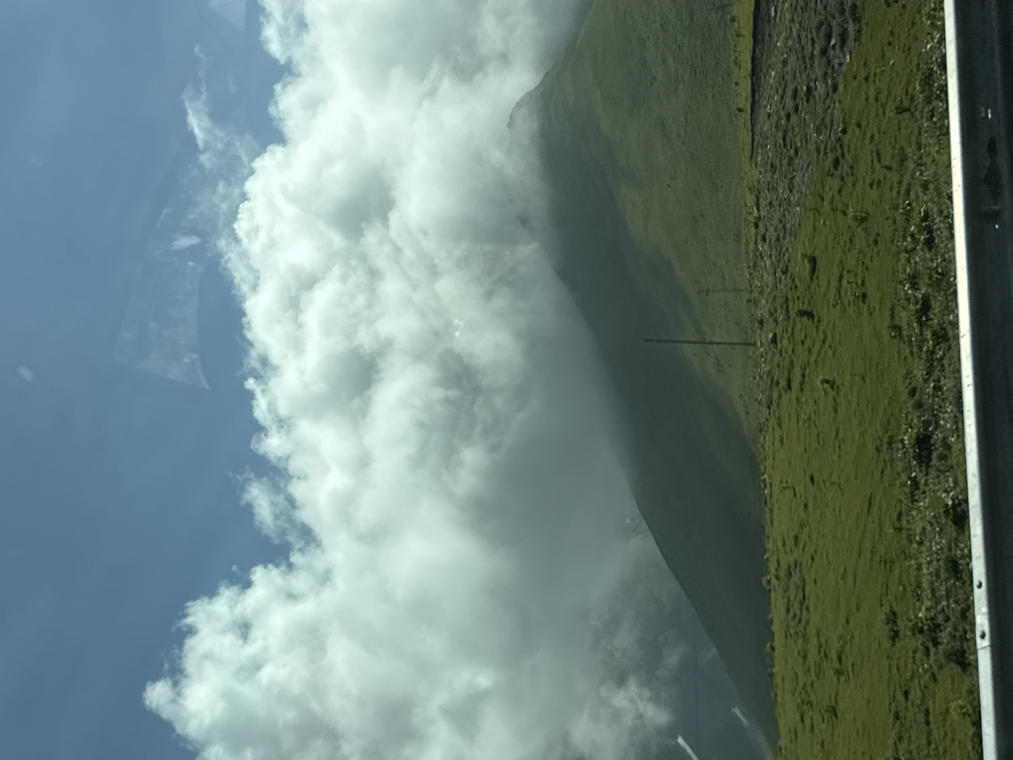 This image captures a scenic view of a mountainous landscape in Gudauri, Georgia. A lush green grassy slope occupies the foreground, leading up to a larger, undulating green hill in the midground. The peak of this hill is partially obscured by thick, white clouds that also drift across the bright blue sky. A tall, thin pole is visible on the grassy slope, and a metal barrier runs along the bottom of the frame. The scene appears to be captured from a moving vehicle, possibly a car, due to reflections visible on the glass in the upper portion of the image. The overall impression is one of natural beauty and open, expansive terrain characteristic of a mountain resort.